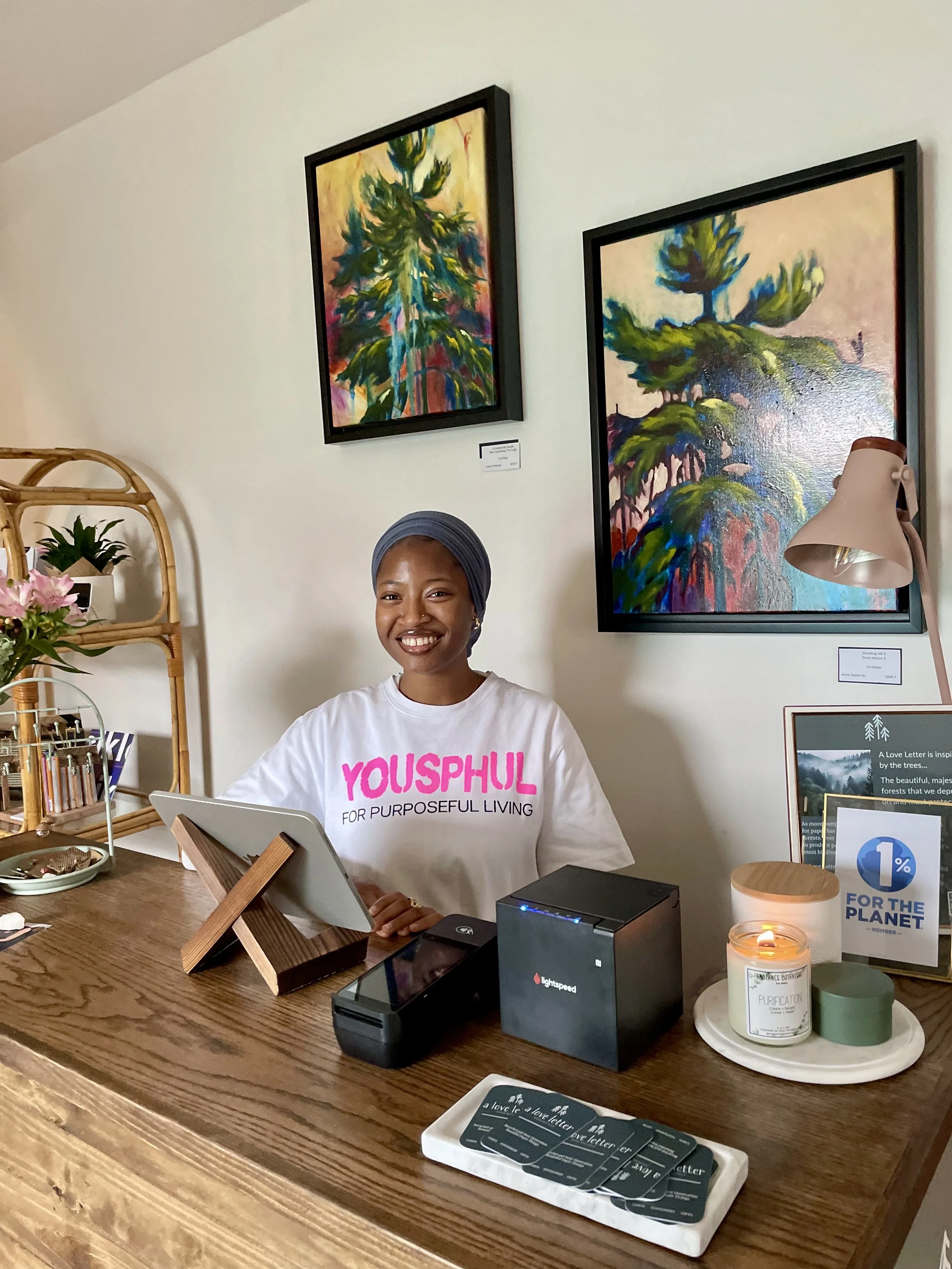 A woman sitting at a wooden desk with a tablet, smiling, wearing a white T-shirt with pink text, and a gray head covering. Behind her are two framed colorful abstract paintings of trees, and on her desk are a candle, a small potted plant, and various
