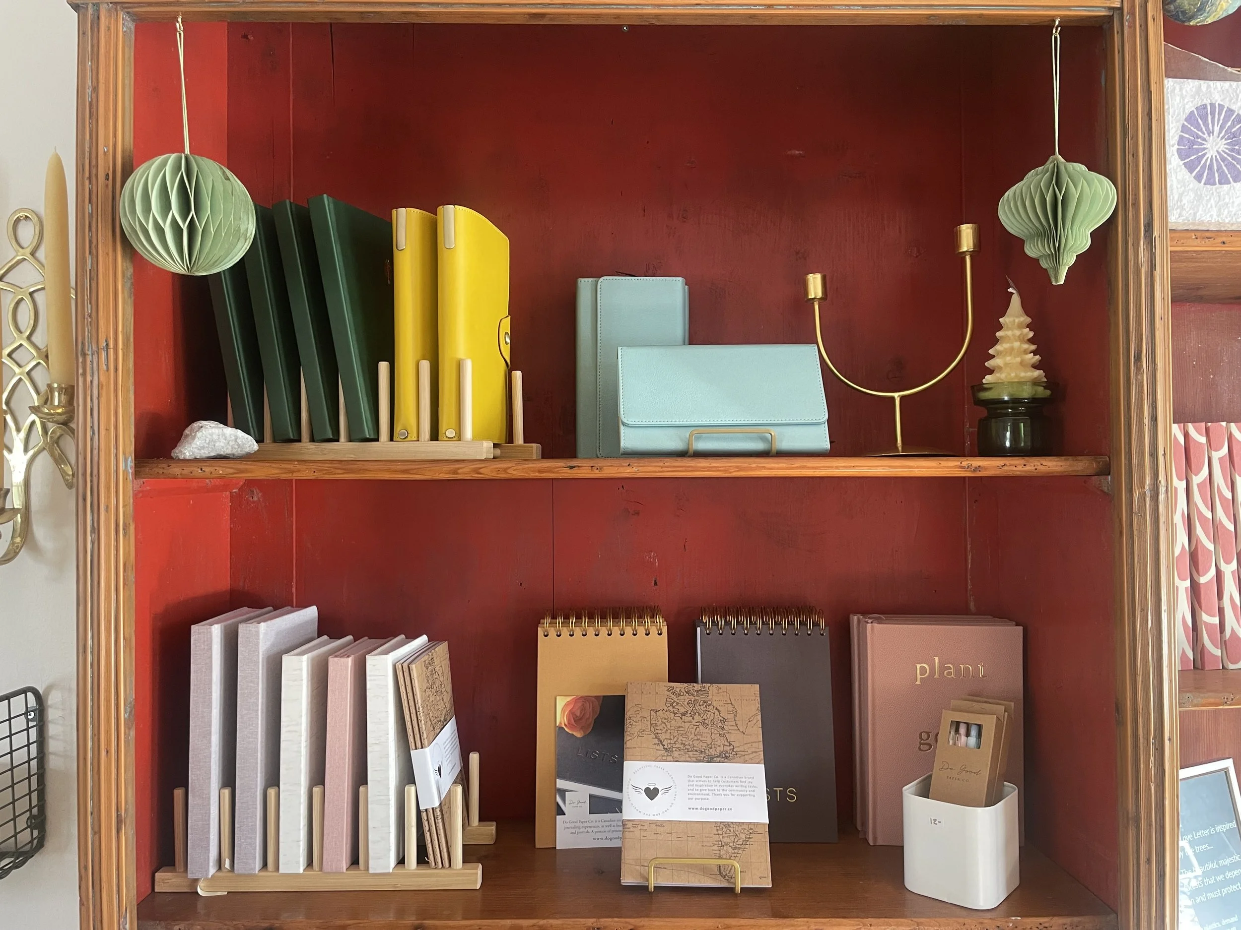 A wooden bookshelf with a red interior and decorated with various colorful items including green and yellow file folders, light blue notebooks, a gold candle holder, a decorative lamp, notebooks, journals, and a small framed picture.