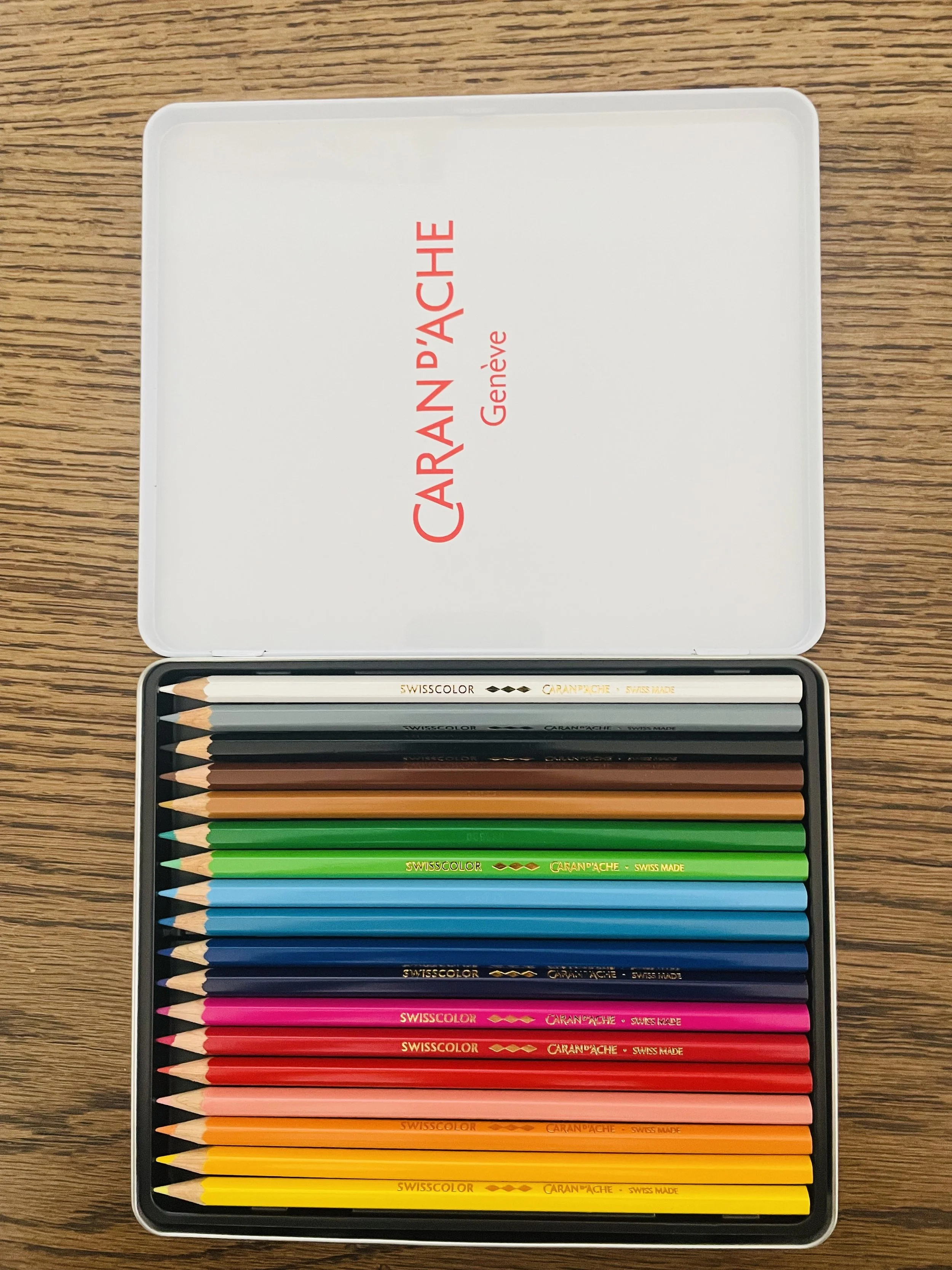 A set of colored pencils in a metal case on a wooden surface. The case lid is open, showing the brand name Caran D'Ache in red and the words 'Genève' on the inside.