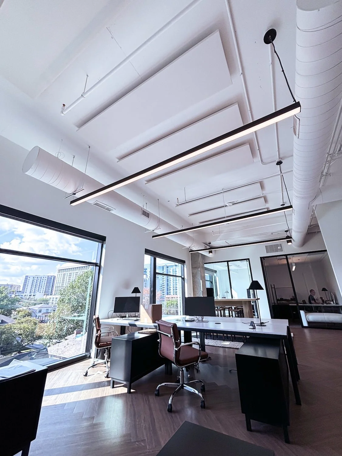 Modern office space with desks, chairs, computers, large windows, and hanging ceiling lights.