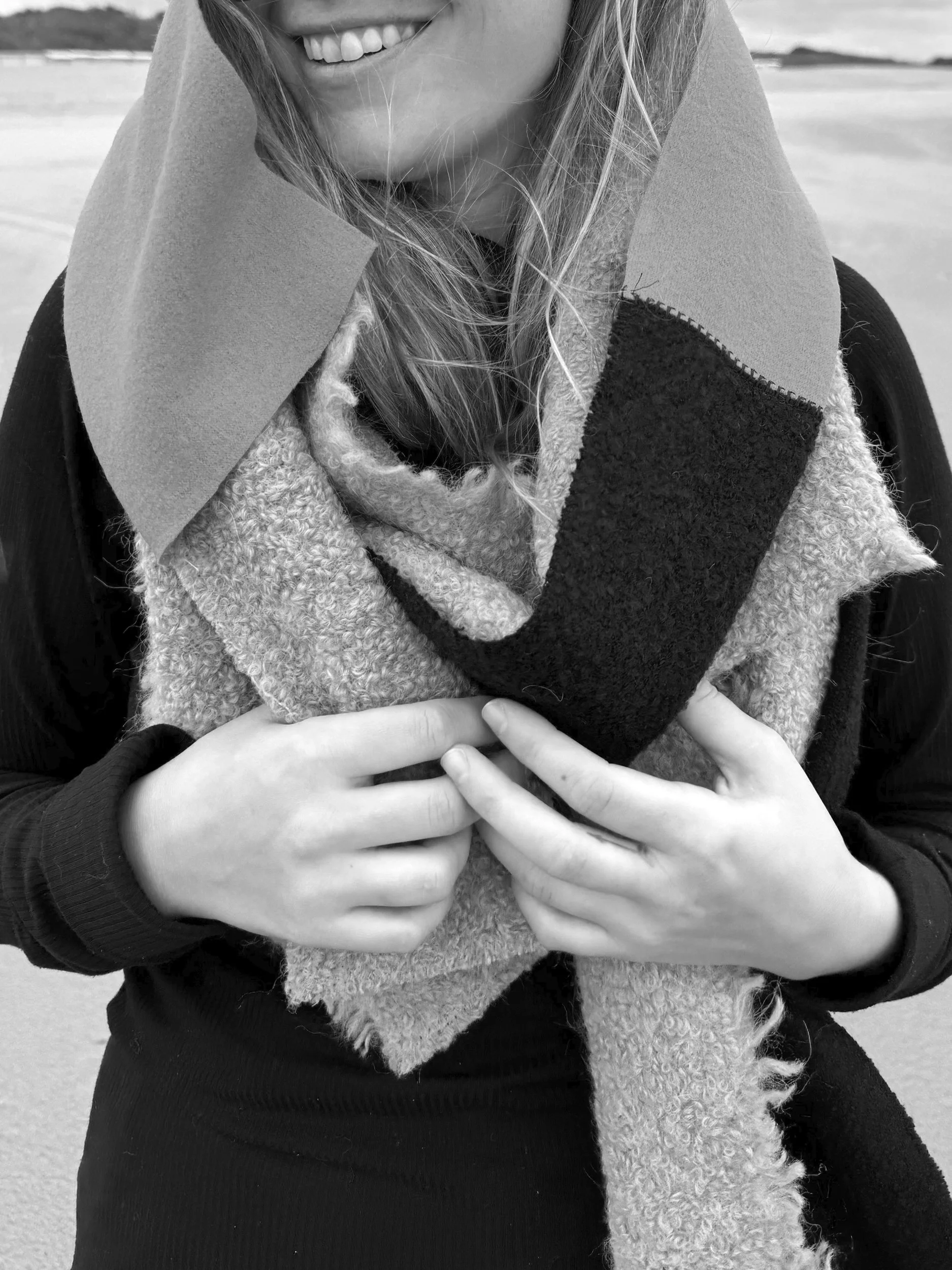 Close-up van een glimlachende vrouw die een kleurrijke sjaal draagt, met hand die haar blouse vasthoudt op de achtergrond van een strand.