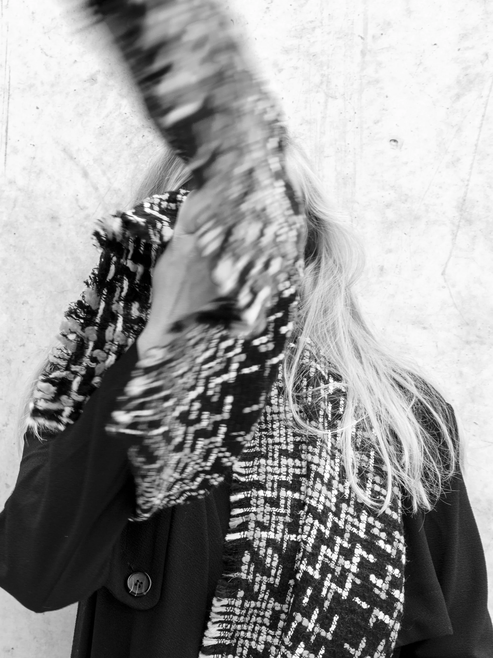 Zwarte en witte gespiegelde jas en haar bedekt het gezicht van een persoon die een geblokte sjaal draagt, met een witte muur op de achtergrond.