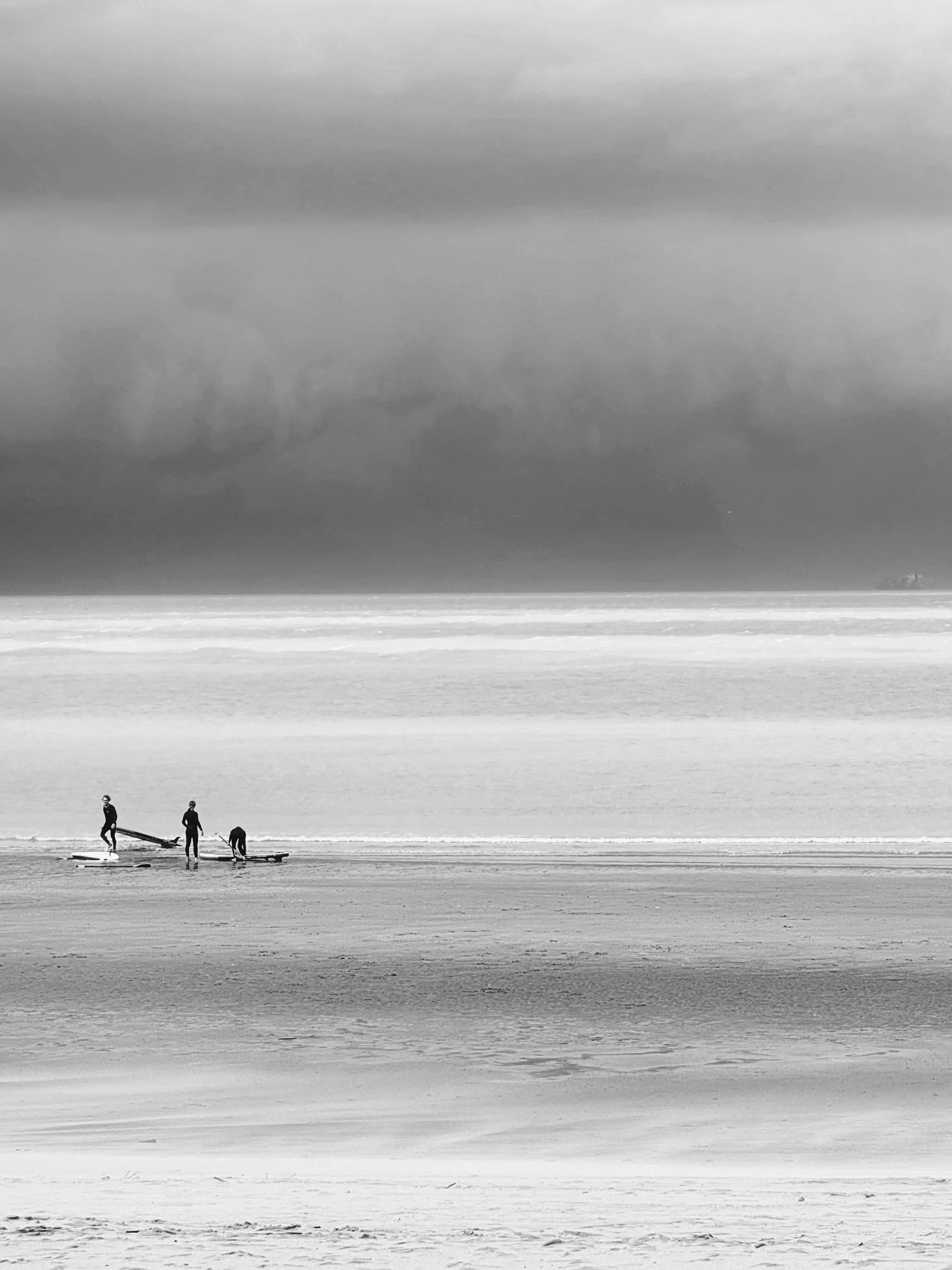 Drie mensen die surfen oefenen op het strand onder een donkere, bewolkte hemel, zwart-wit foto.