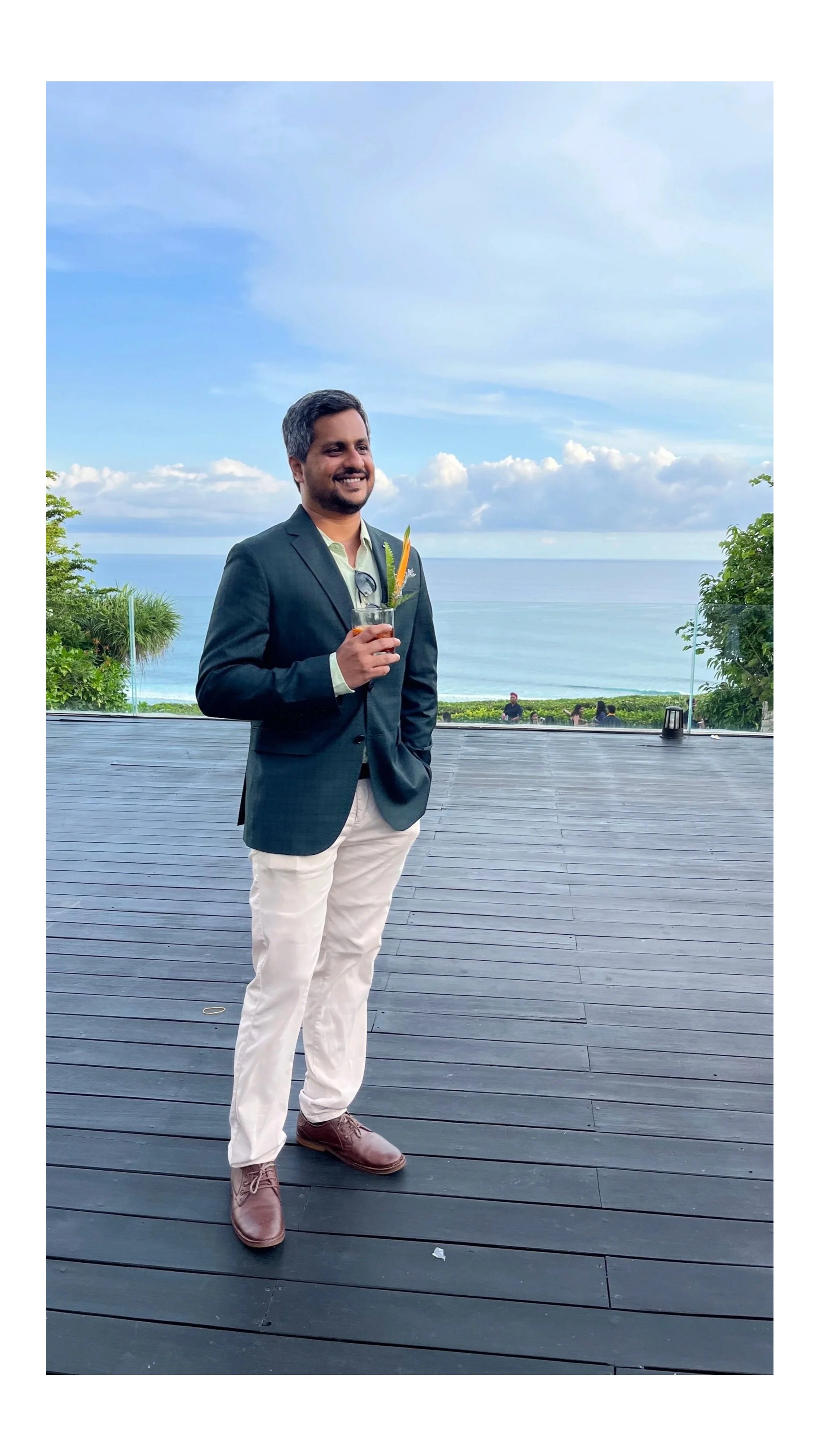 A man dressed in a navy blazer, light green shirt, and khaki pants holding a cocktail, standing outdoors on a wooden deck with a view of the ocean and cloudy sky in the background.