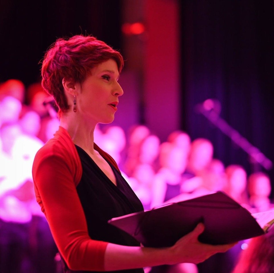Catriona Bühler Sopran mit kurzen roten Haaren, die in einem dunklen Raum bei pinker Beleuchtung einen Chor leitet.