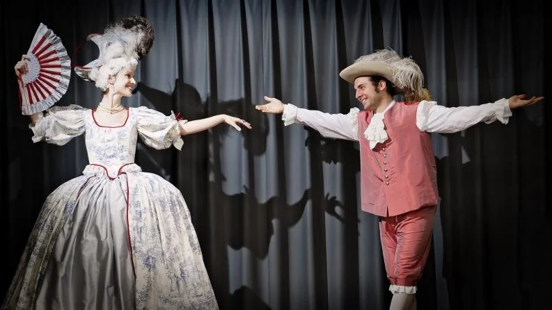 Zwei Menschen in historische Kostüme auf einer Bühne, die sich gegenseitig zu begrüßen scheinen, vor einem schwarzen Vorhang.