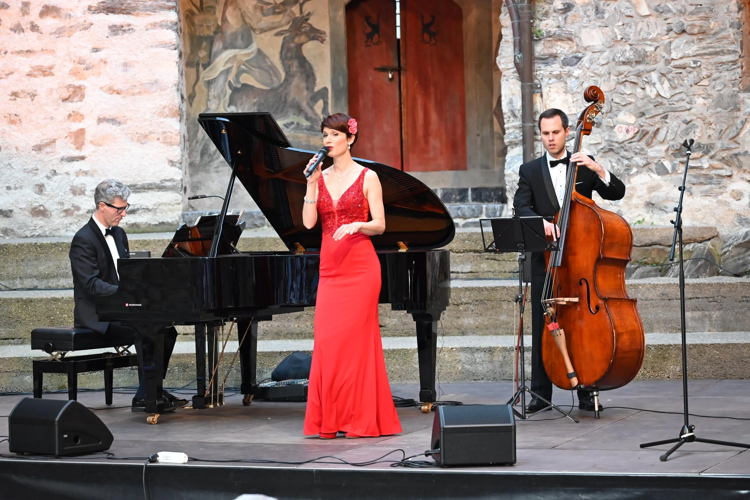 Catriona Bühler, Sopran mit Samuel Zünd, Klavier und Fridolin Blumer, Kontrabass, und einem dunklen Kronleuchter im Hintergrund, die auf einer historischen Bühne mit Steinmauer im Freien auftreten.