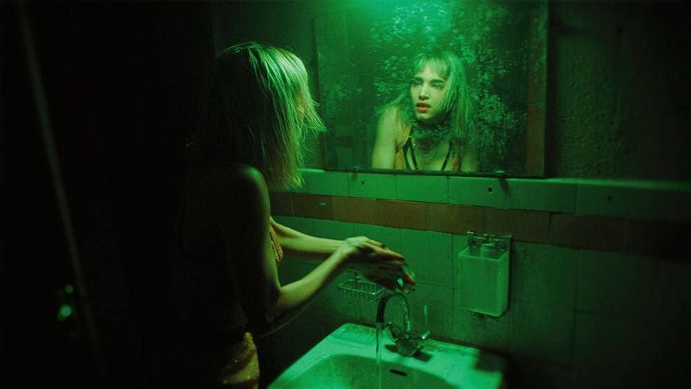 Une femme blonde se regarde dans un miroir entouré d'une lumière verte dans une salle de bain, avec un mur en tuiles derrière, et une peau sombre derrière elle dans le reflet.