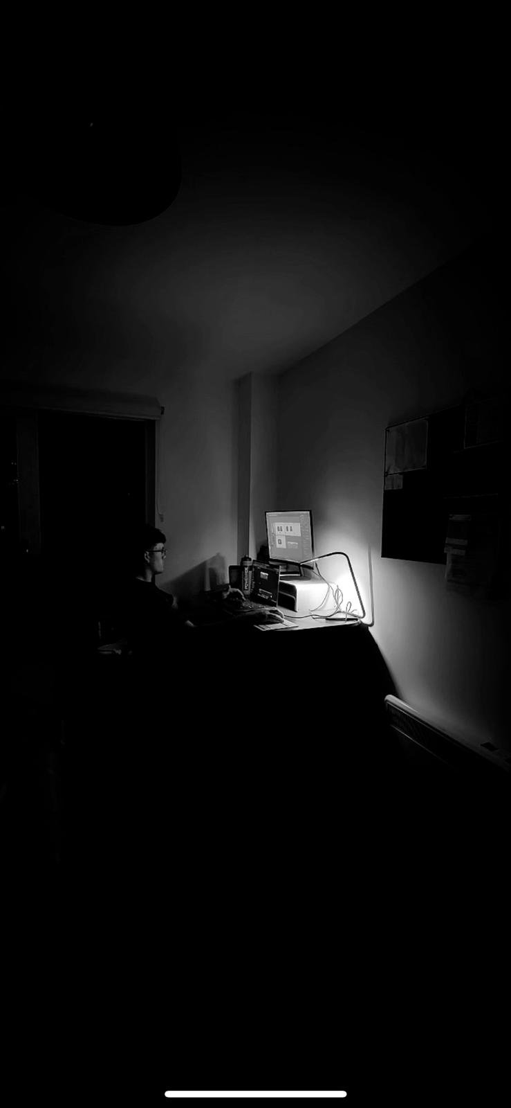 A person sitting at a desk in a dimly lit room, working on a computer with a bright monitor.