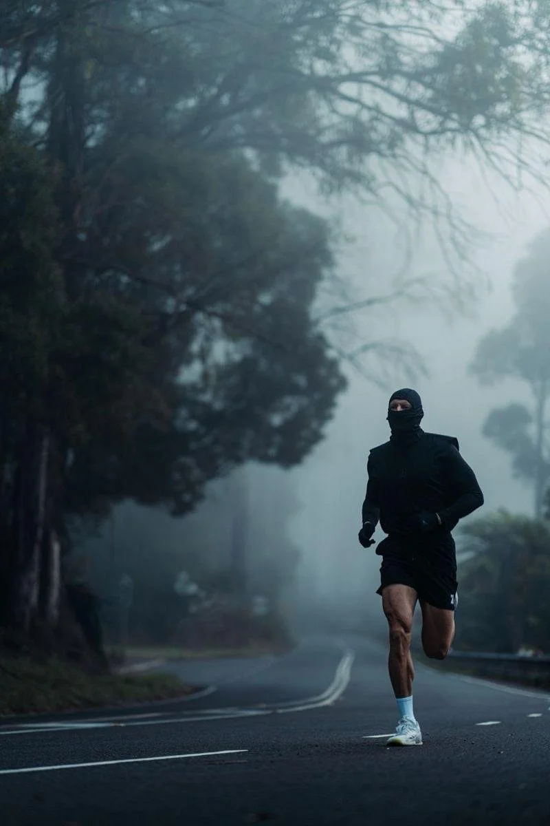 A person running on a foggy, winding road surrounded by trees, dressed in dark athletic gear with a face covering.