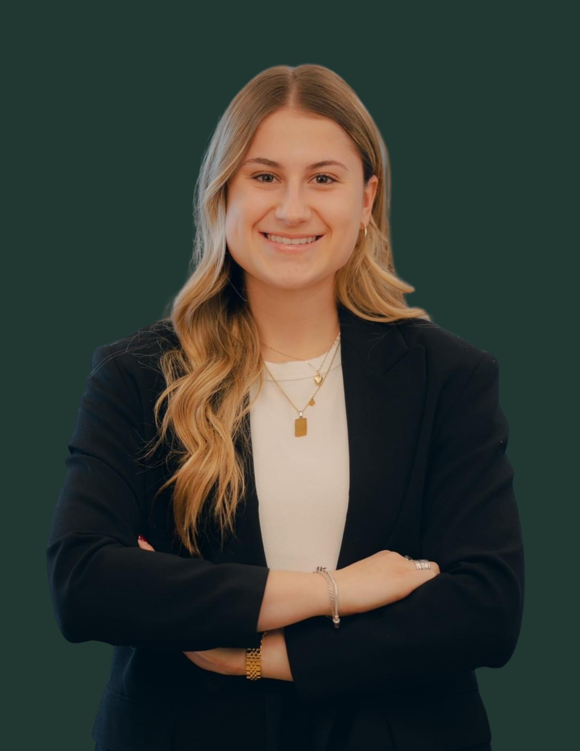 Une jeune femme souriante avec des cheveux longs et ondulés, portant une veste noire, des bijoux en or, et une chemise blanche, posant avec les bras croisés devant un fond vert foncé.