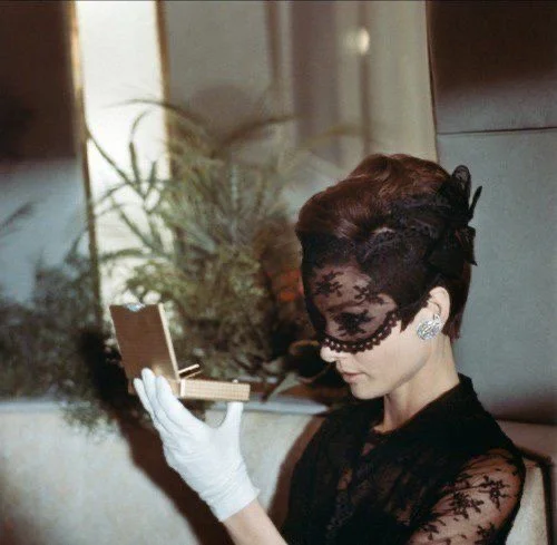 A woman with short Hairstyle wearing a black lace mask and gloves, looking at a compact mirror, seated indoors with plants in the background.