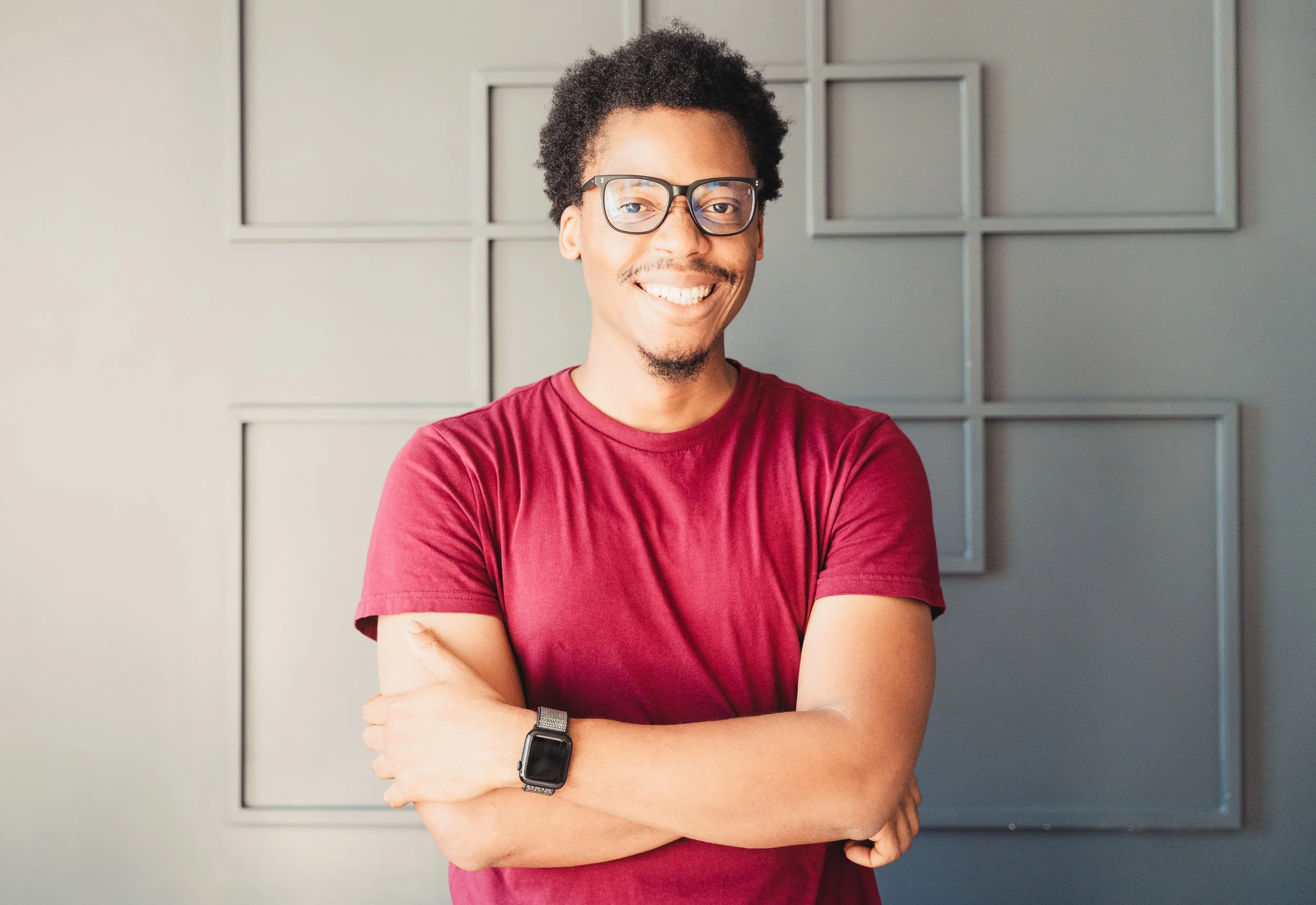 A smiling man with glasses and a watch, wearing a red T-shirt, standing with arms crossed in front of a gray wall with decorative geometric panels.