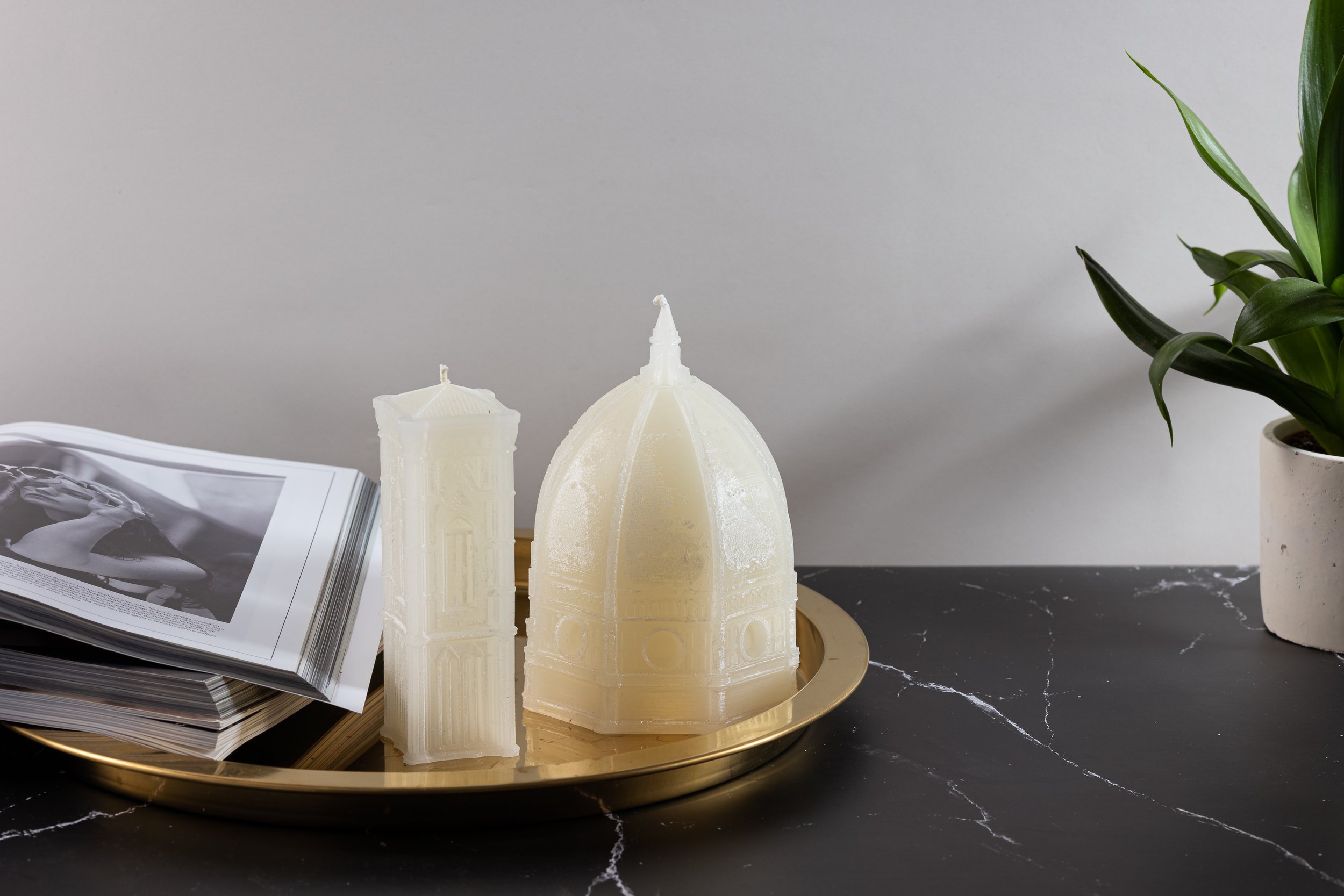 Decorative candle shaped like a dome building and a tower on a gold tray, with an open magazine, and a potted plant on a black marble surface against a plain light-colored wall.