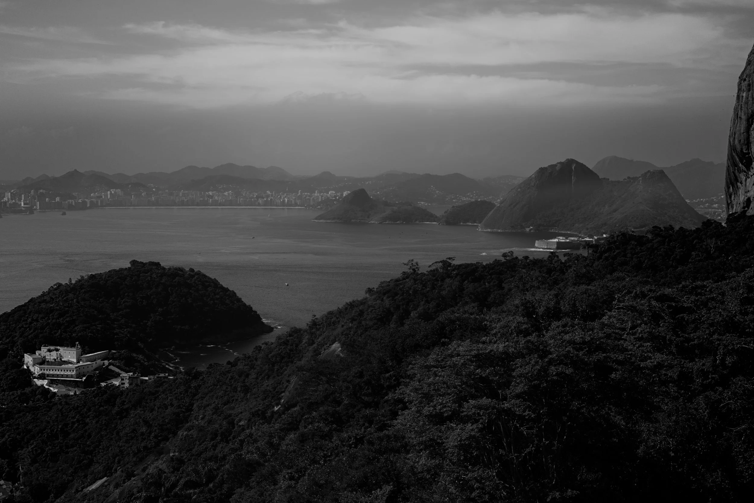 A black and white landscape of a bay with mountains in the background, hills in the foreground, and a large cliff on the right side.