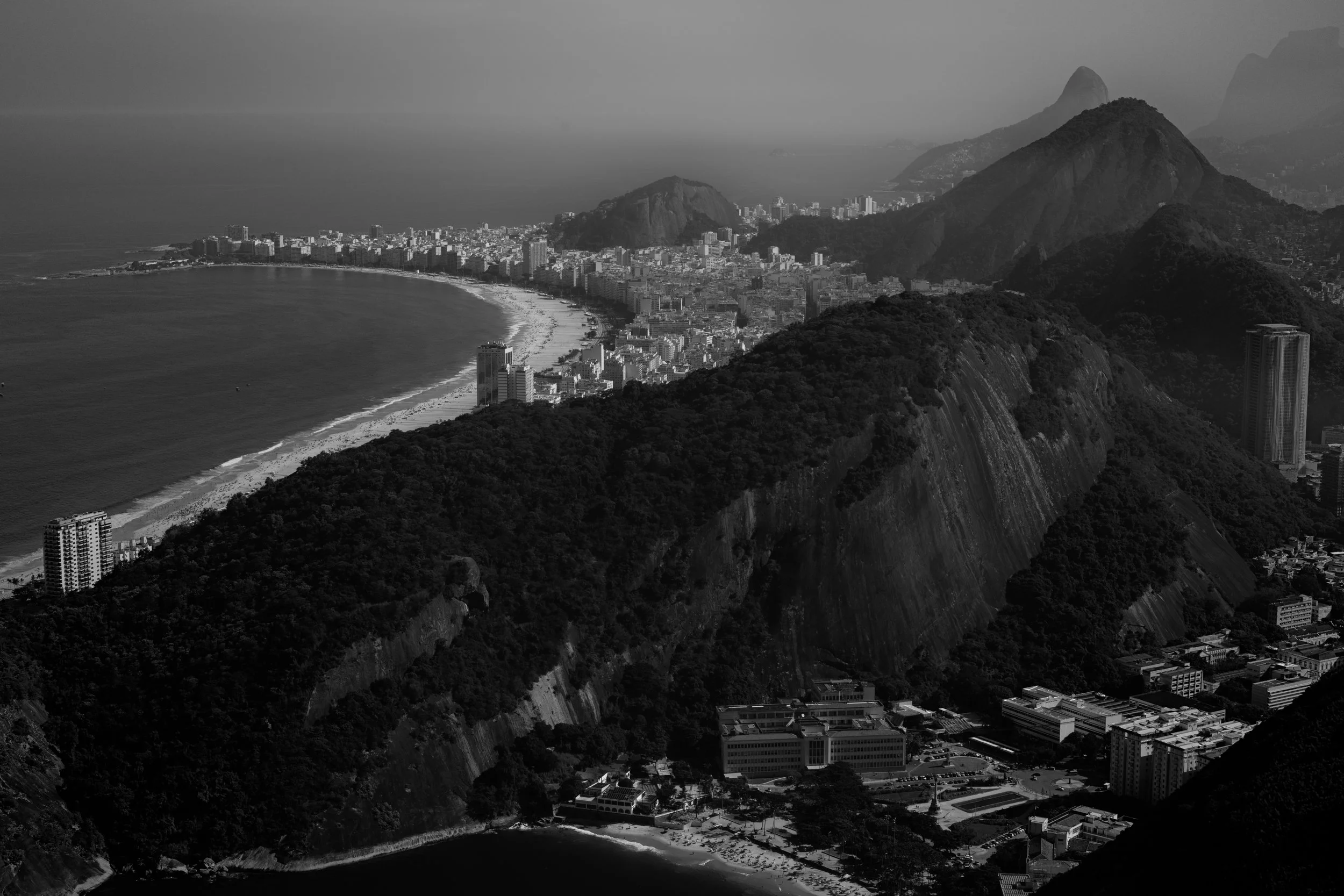 A black and white aerial view of a coastal city with a beach, high-rise buildings, and surrounding mountains.