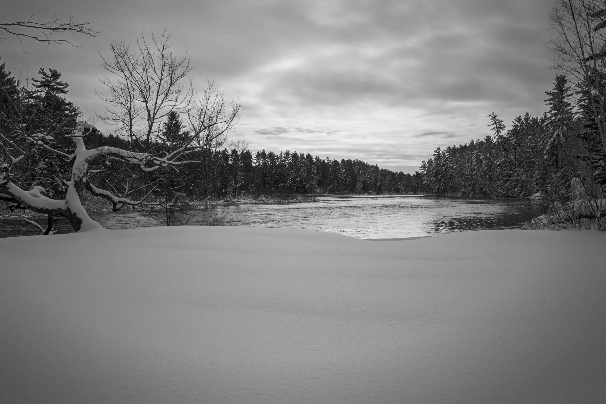 Black and white photograph of a winter scene with snow-covered ground, leafless trees, a river, and a cloudy sky.