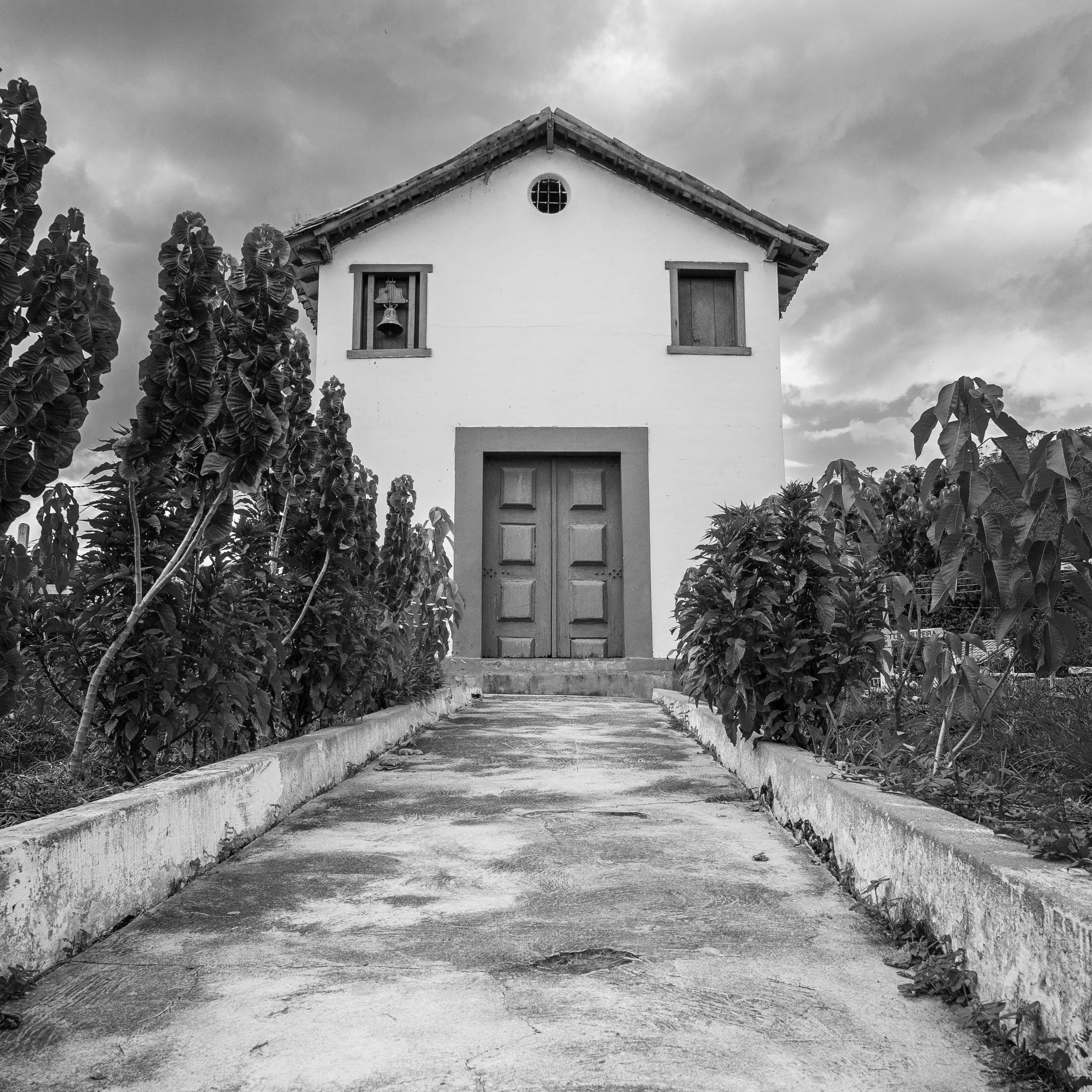 A black-and-white photo of a small Brazilian cappella with a triangular roof, a small circular window, and a bell on the front wall. The house is surrounded by dense foliage and trees, with a cloudy sky above.