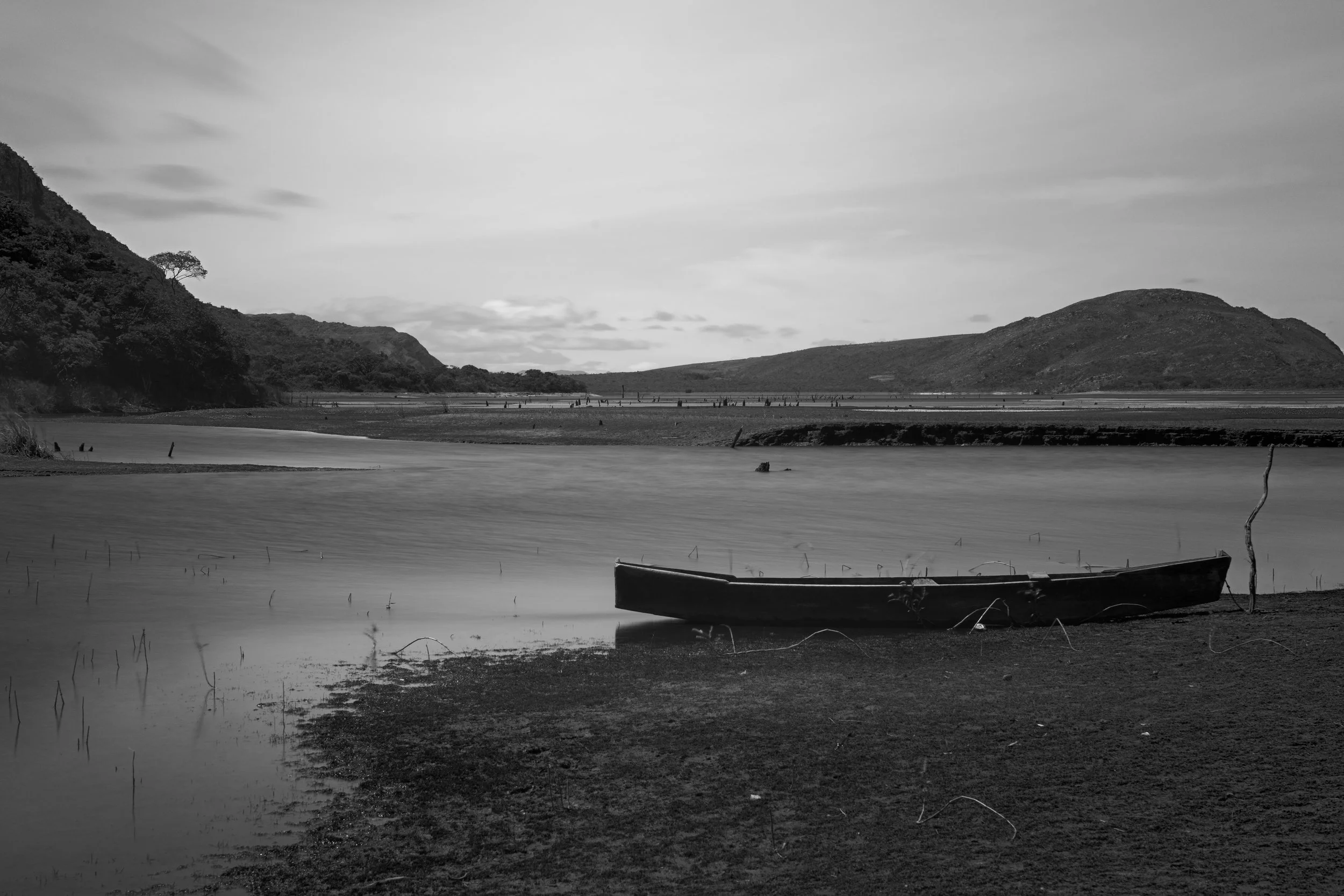 A black and white landscape photograph of a river or lake with a small boat resting on the shore in the foreground, surrounding hills or mountains, and a cloudy sky.