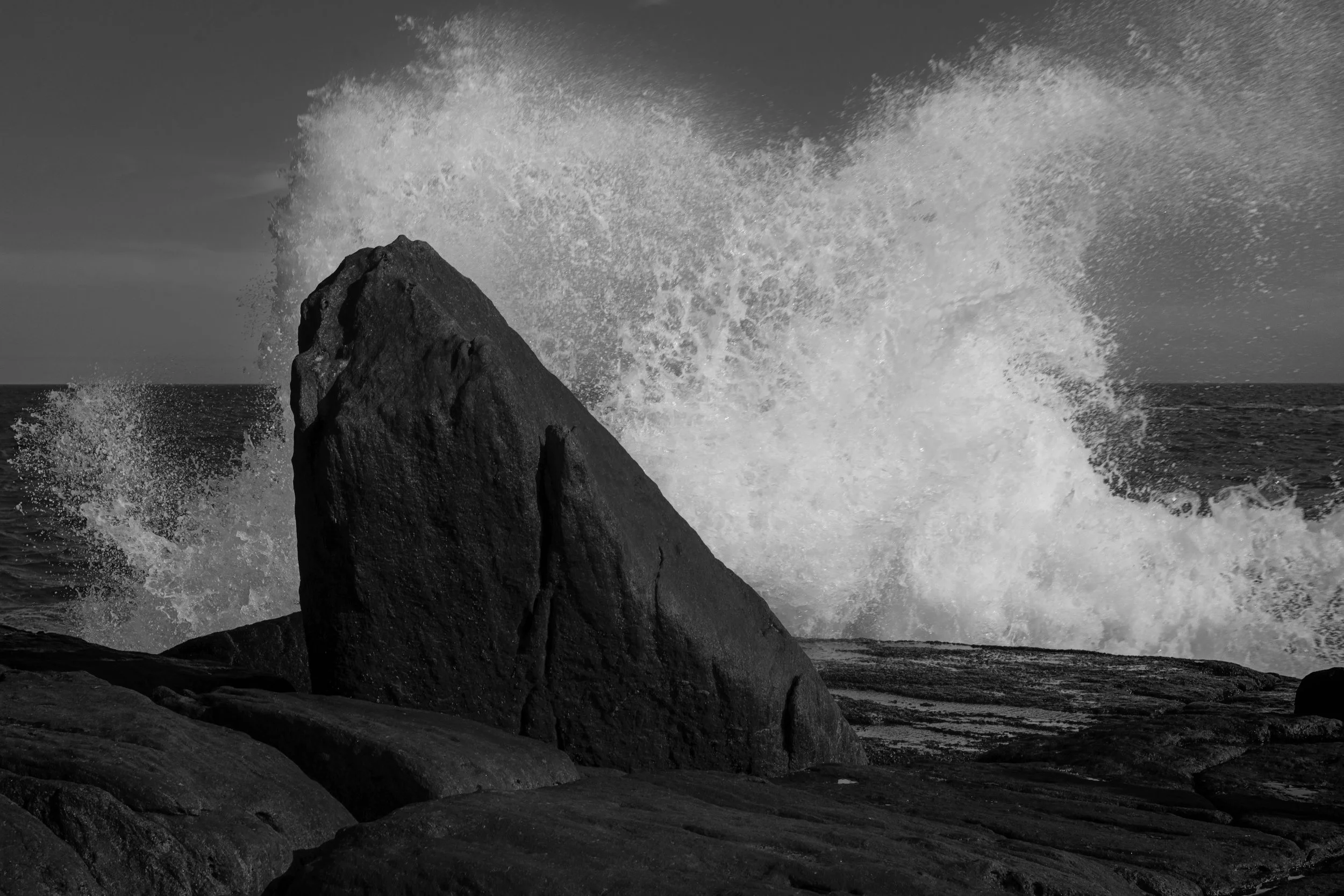 Black and white photograph of waves crashing against large rocks along the shoreline.