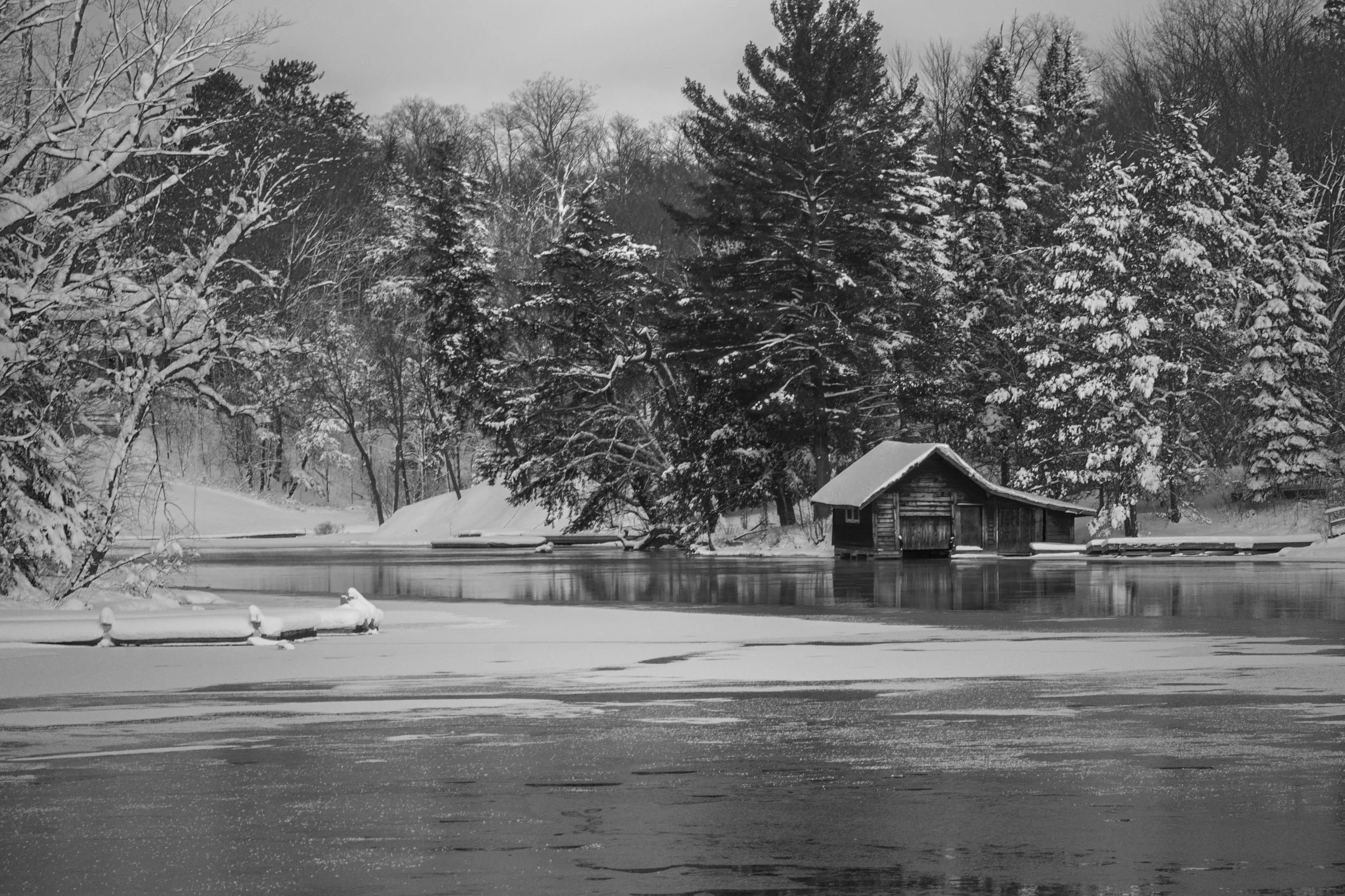 A peaceful winter scene with snow-covered trees and a small cabin near a frozen lake, black and white photograph.