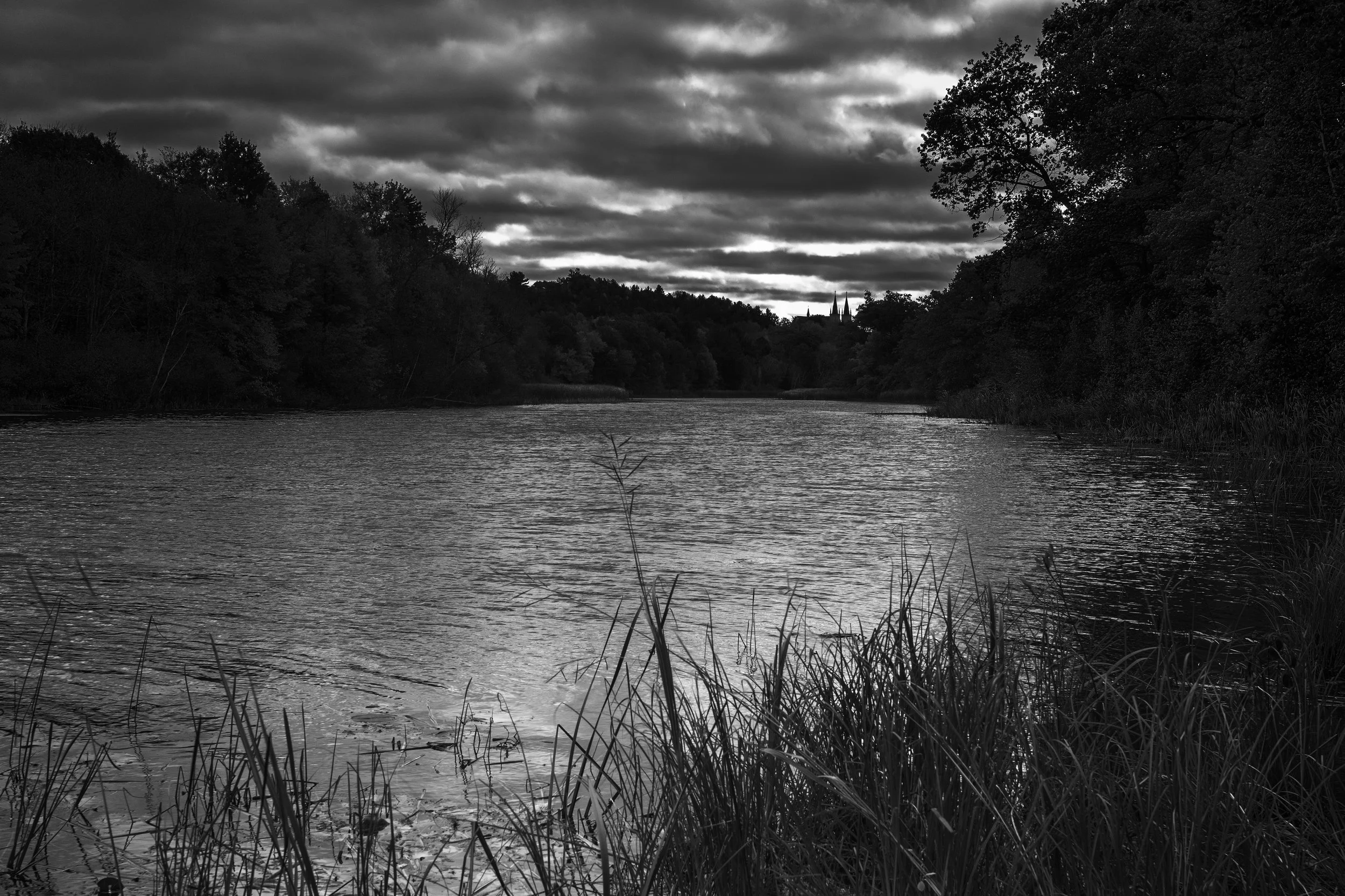 A black and white photo of a river lined with trees, with dark clouded sky overhead and a distant church with tall spires visible in the background.