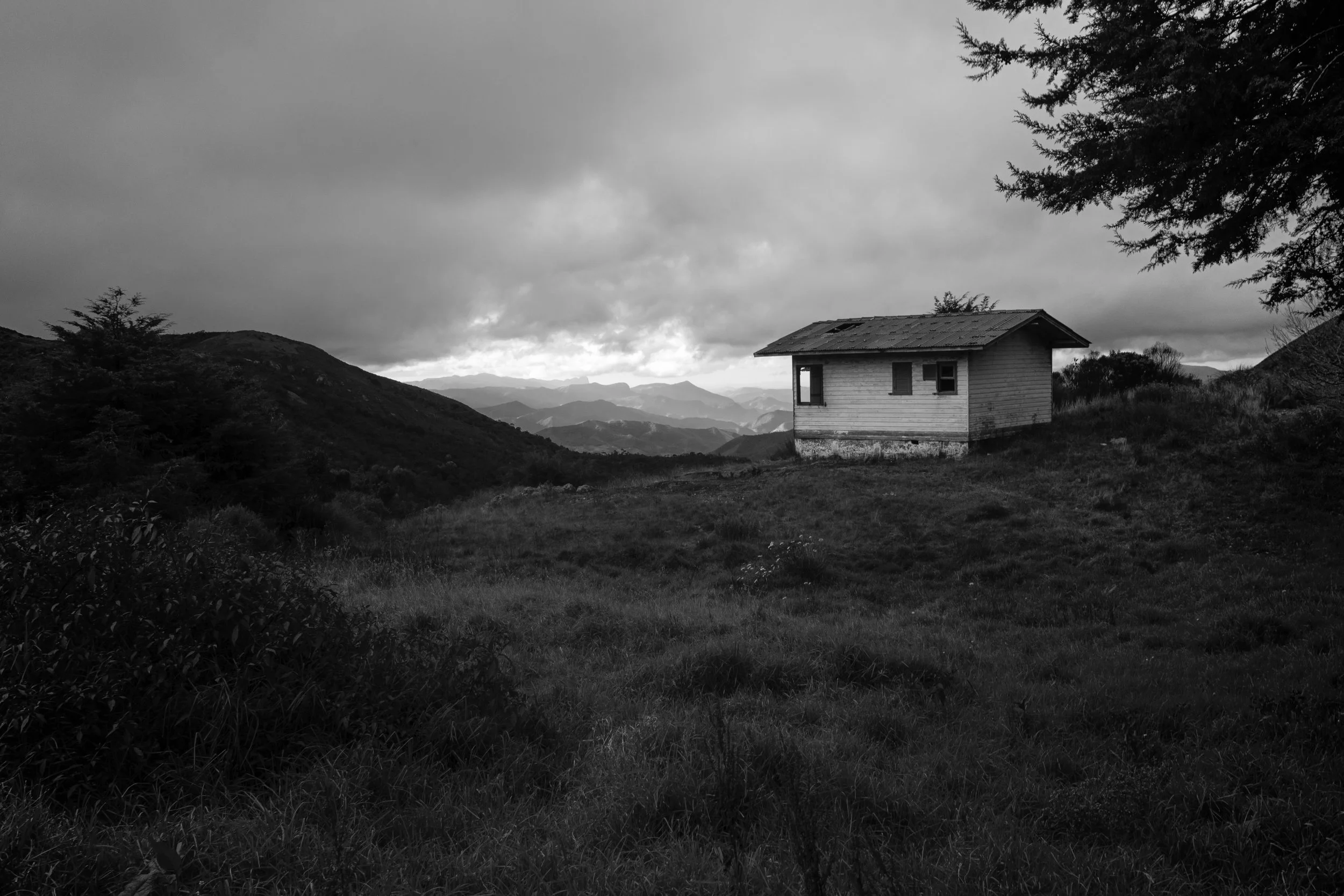 A small wooden house with a sloped roof situated on a hill, surrounded by trees and overlooking distant mountain ranges under a cloudy sky.