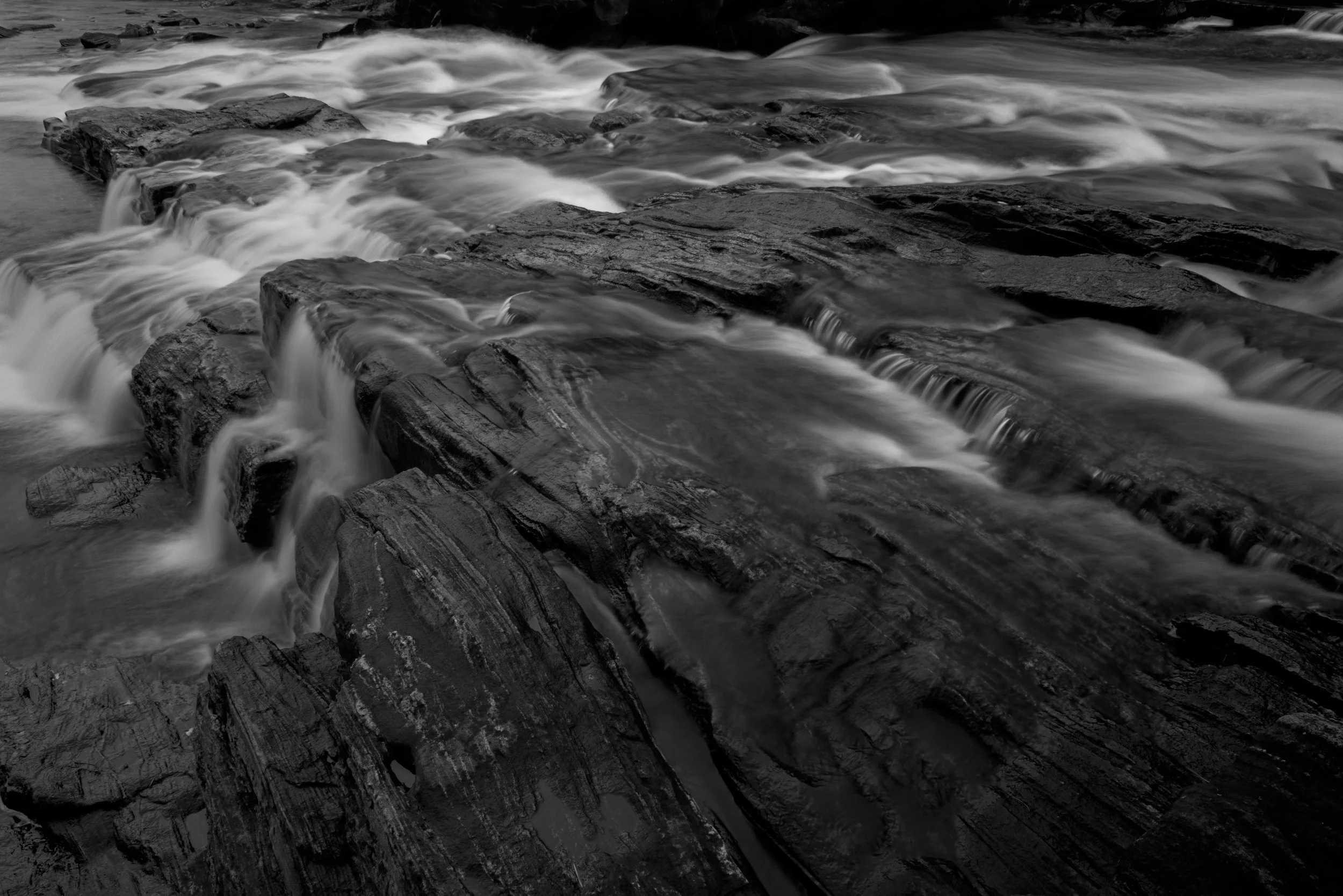 Black and white photograph of flowing water cascading over rocks in a river or stream.