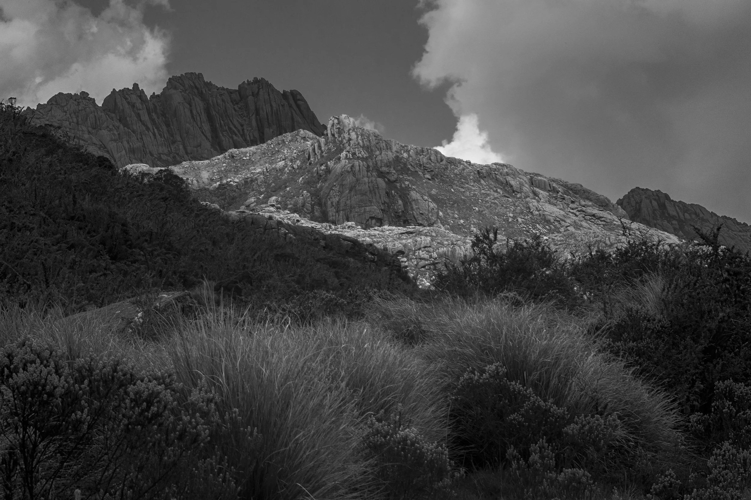Black and white photograph of a rugged mountain range with rocky peaks and some smoke or steam rising from the top, framed by grass and bushes in the foreground.