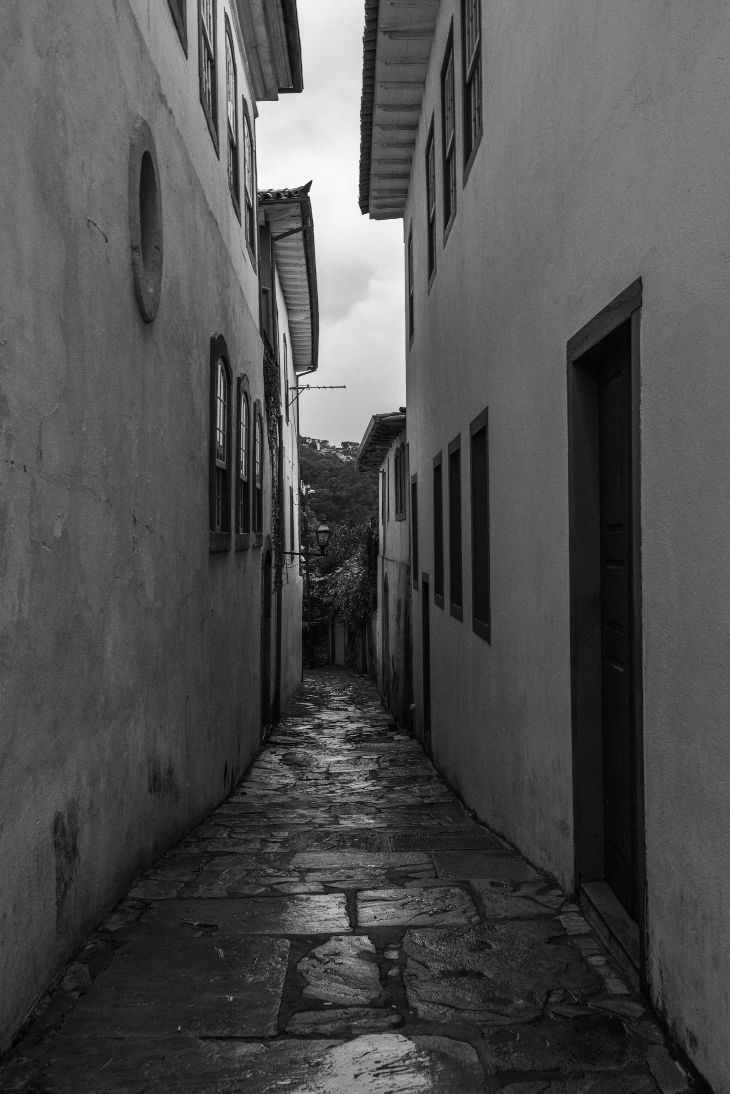 A narrow old cobblestone alley between two tall buildings with traditional windows, leading to a distant view of hills and trees, and an overcast sky.