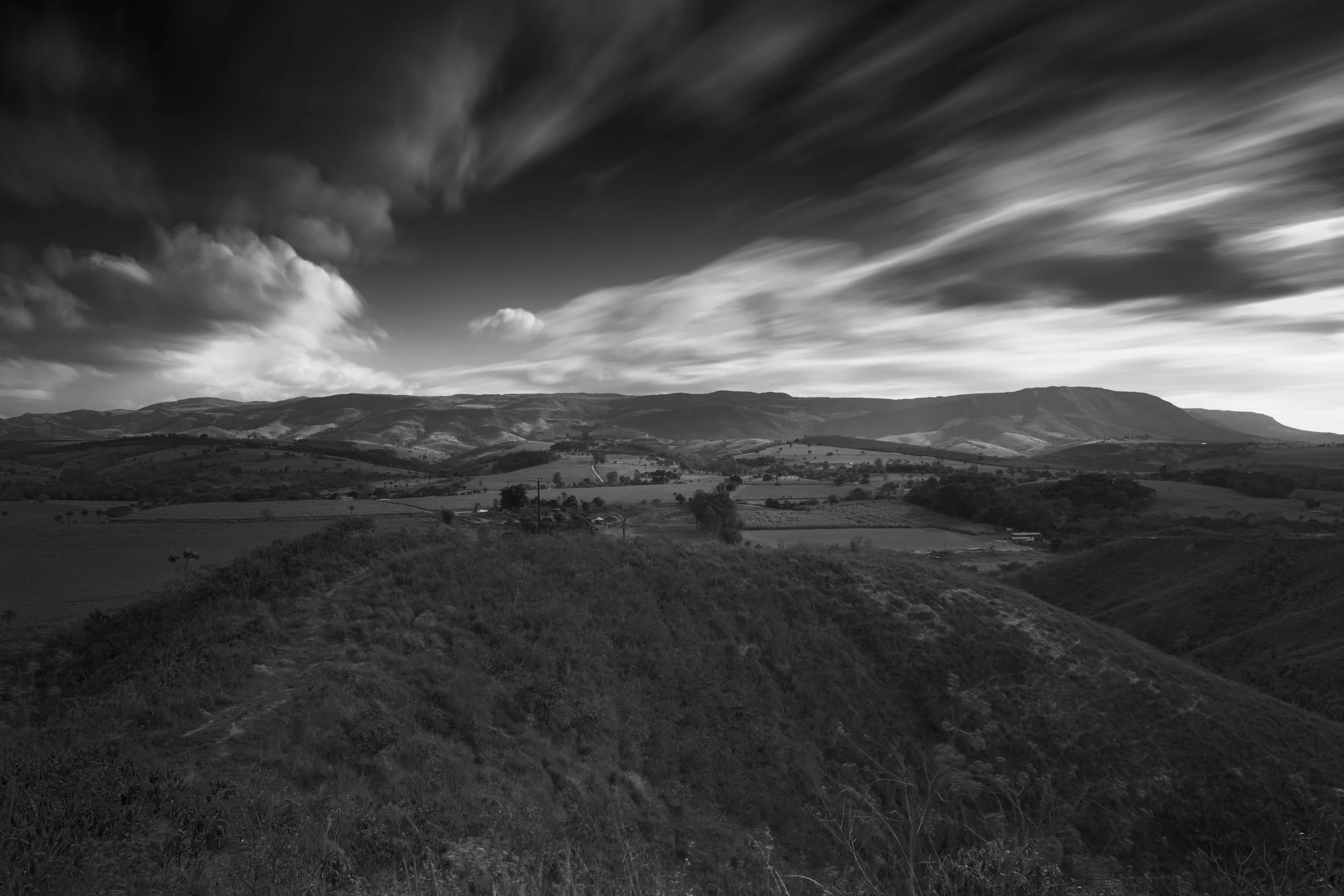 A black-and-white landscape photograph of rolling hills and mountains under a sky filled with wispy clouds.