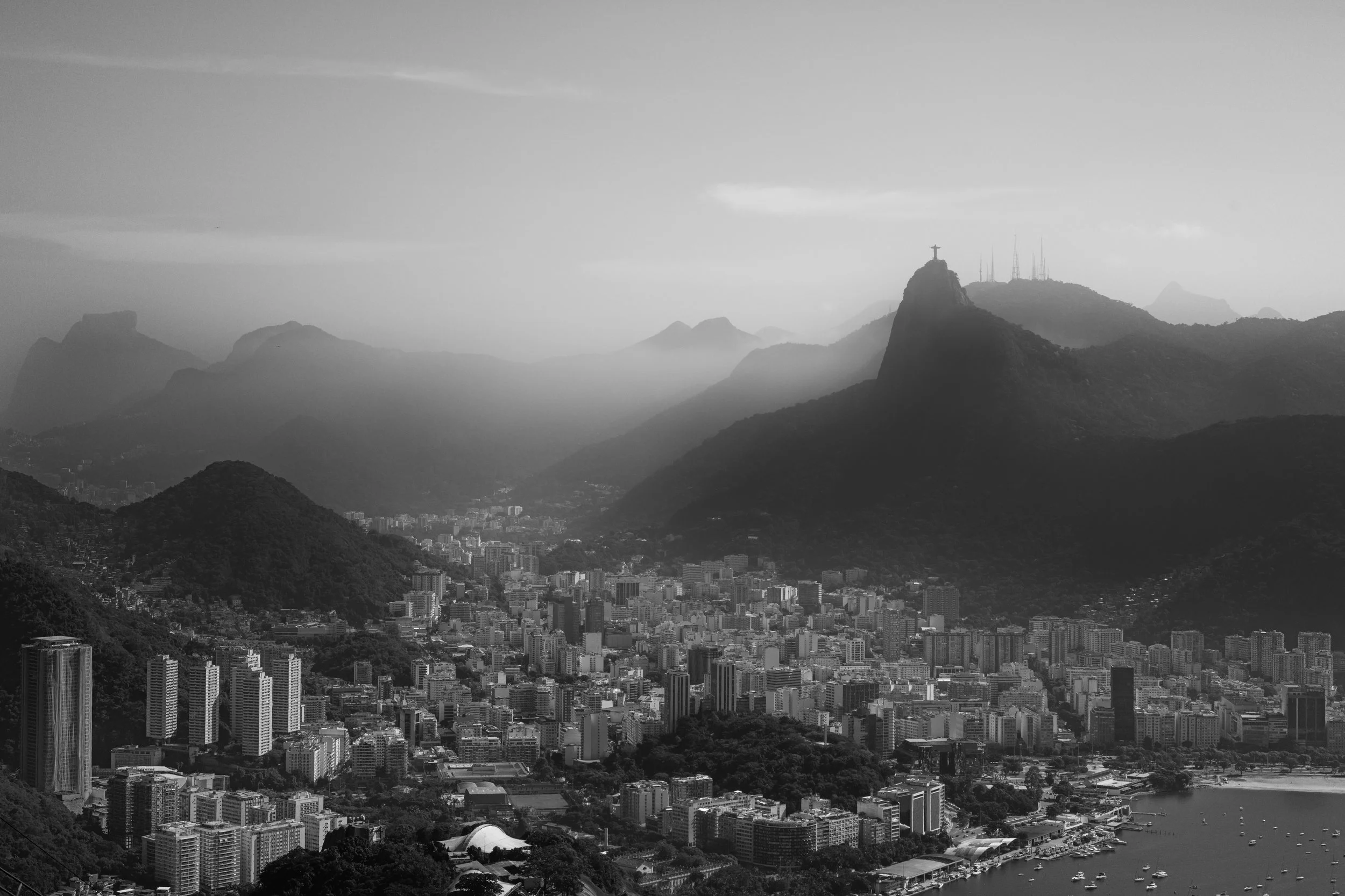 A panoramic view of a city with tall buildings and mountains in the background, including a mountain with the Christ the Redeemer statue on top.