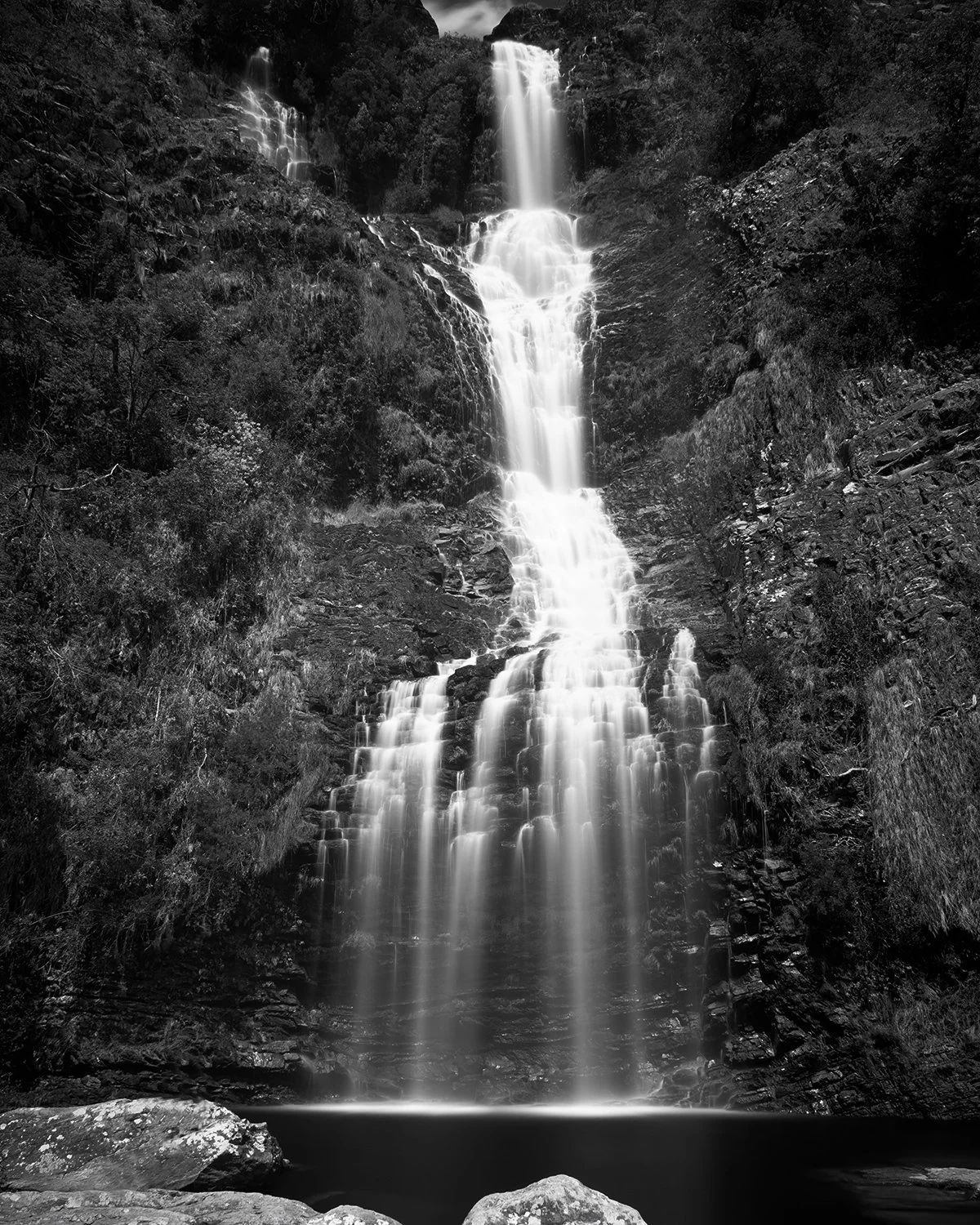 A black and white photograph of a multi-tiered waterfall cascading down a rocky cliffside with surrounding trees and foliage.