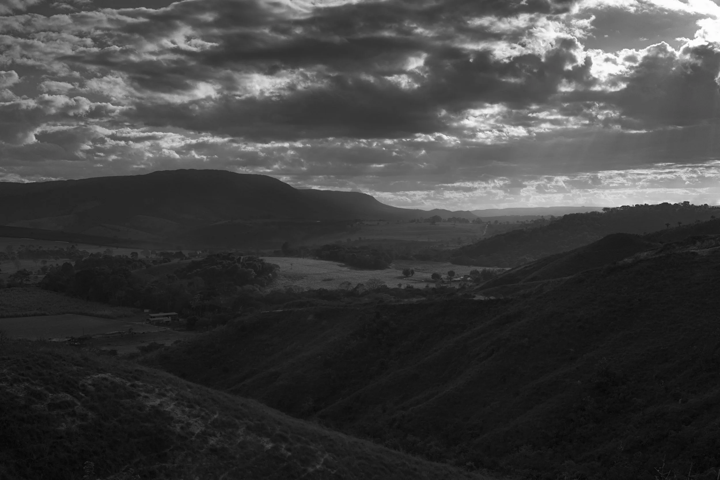 A black and white landscape photograph showing rolling hills and mountains under a dramatic sky filled with clouds.