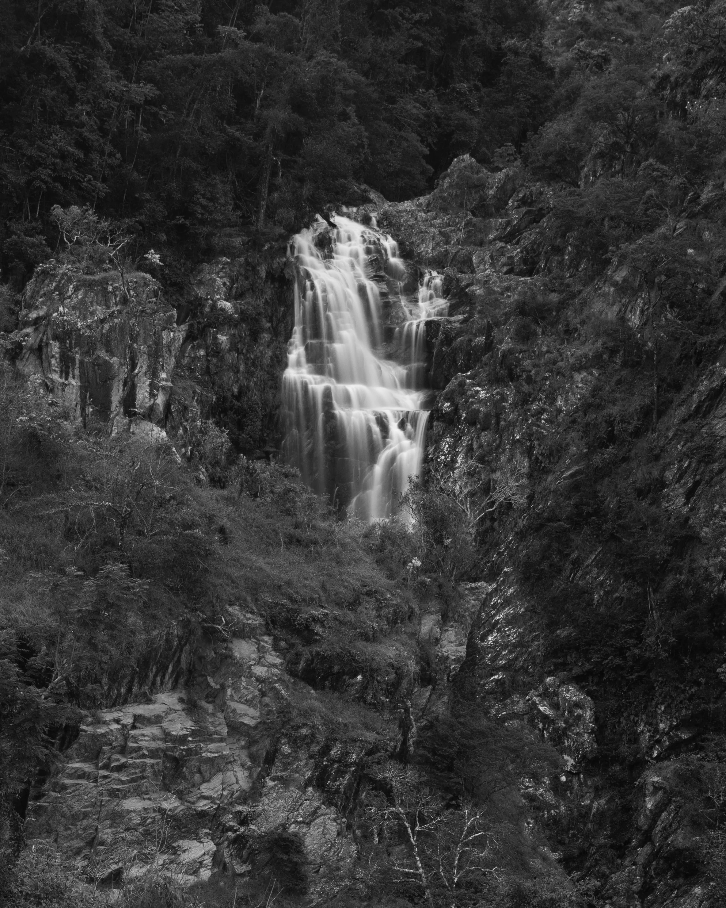 A black-and-white photograph of a multi-tiered waterfall cascading down a rocky hillside surrounded by dense forest.