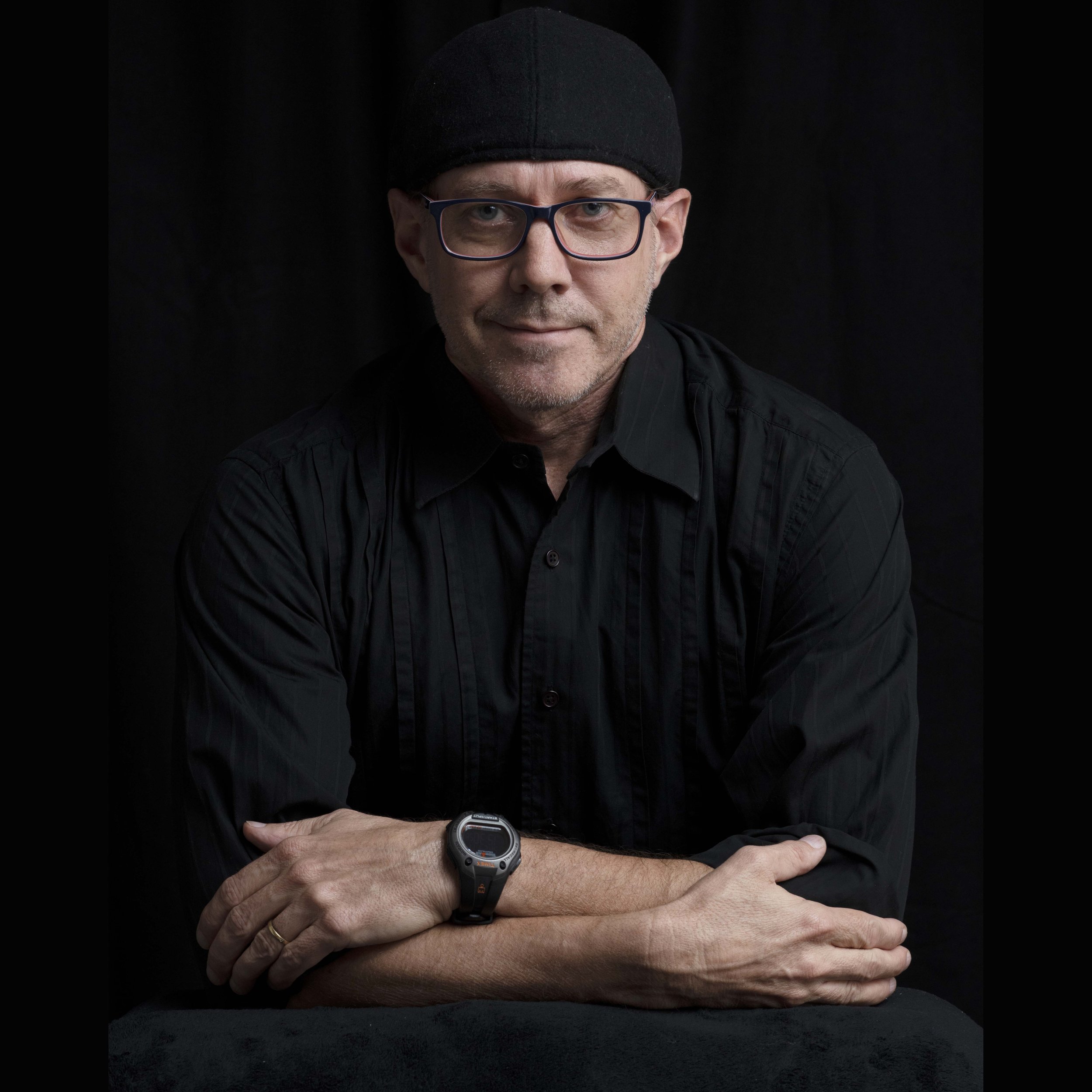 A middle-aged man wearing a black cap, glasses, and a black shirt, resting his arms on a black surface, with a watch on his left wrist, against a black background.