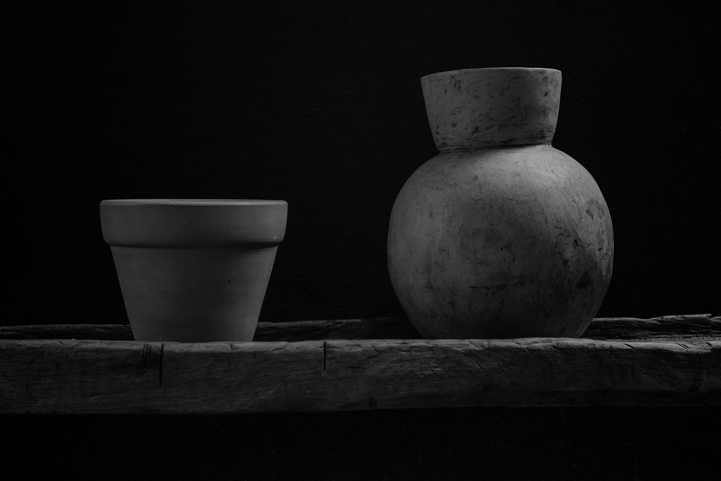 Black and white photo of two pottery vases on a wooden plank with a dark textured background. The larger vase has a rounded body with a narrow opening, while the smaller vase has a wider opening and tapers towards the base.