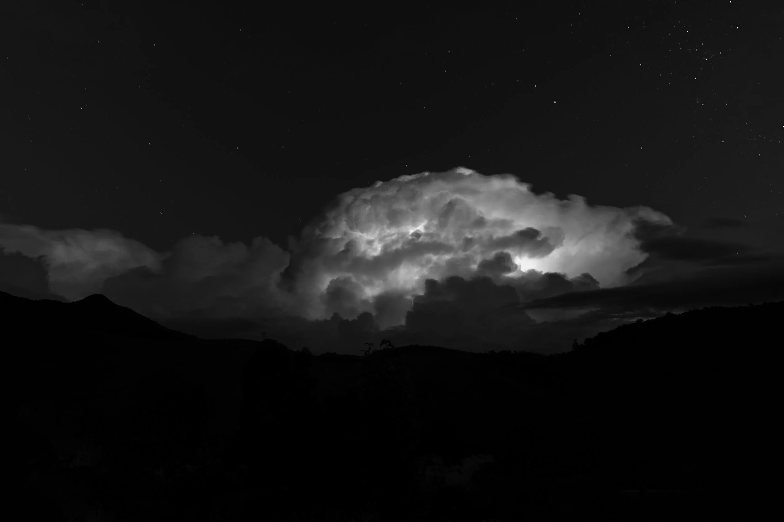 Nighttime landscape with clouds illuminated by lightning and stars in the sky, silhouette of mountains in the foreground.