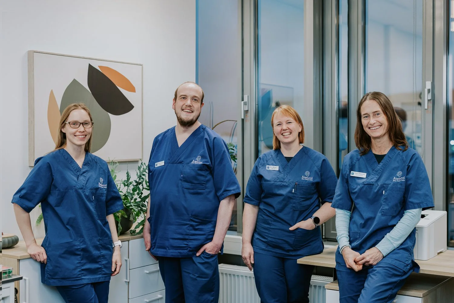 Fire helsepersonell i blå uniformer står og smiler i et rom med store vinduer og moderne innredning.