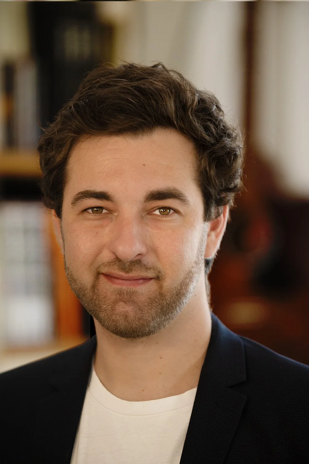 Portrait d'un homme avec cheveux bruns, barbe légère, portant une veste noire et un t-shirt blanc, dans un intérieur chaleureux.