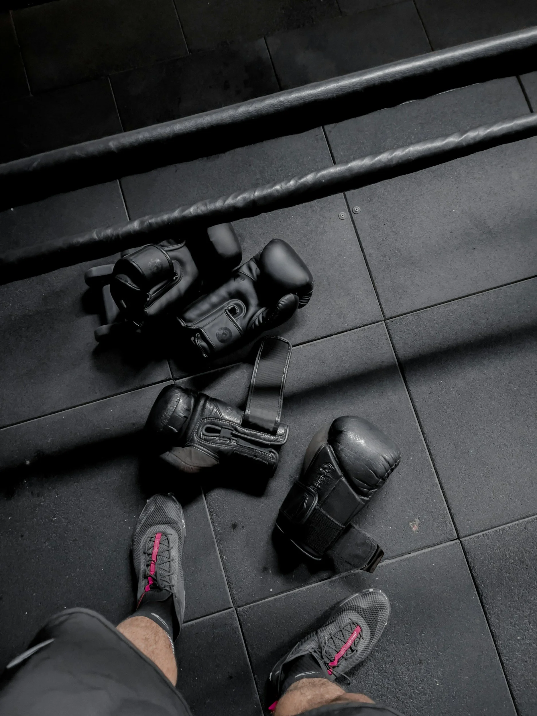 A person in gym shoes stands on black rubber gym floor next to boxing gloves and equipment, with gym railing overhead.