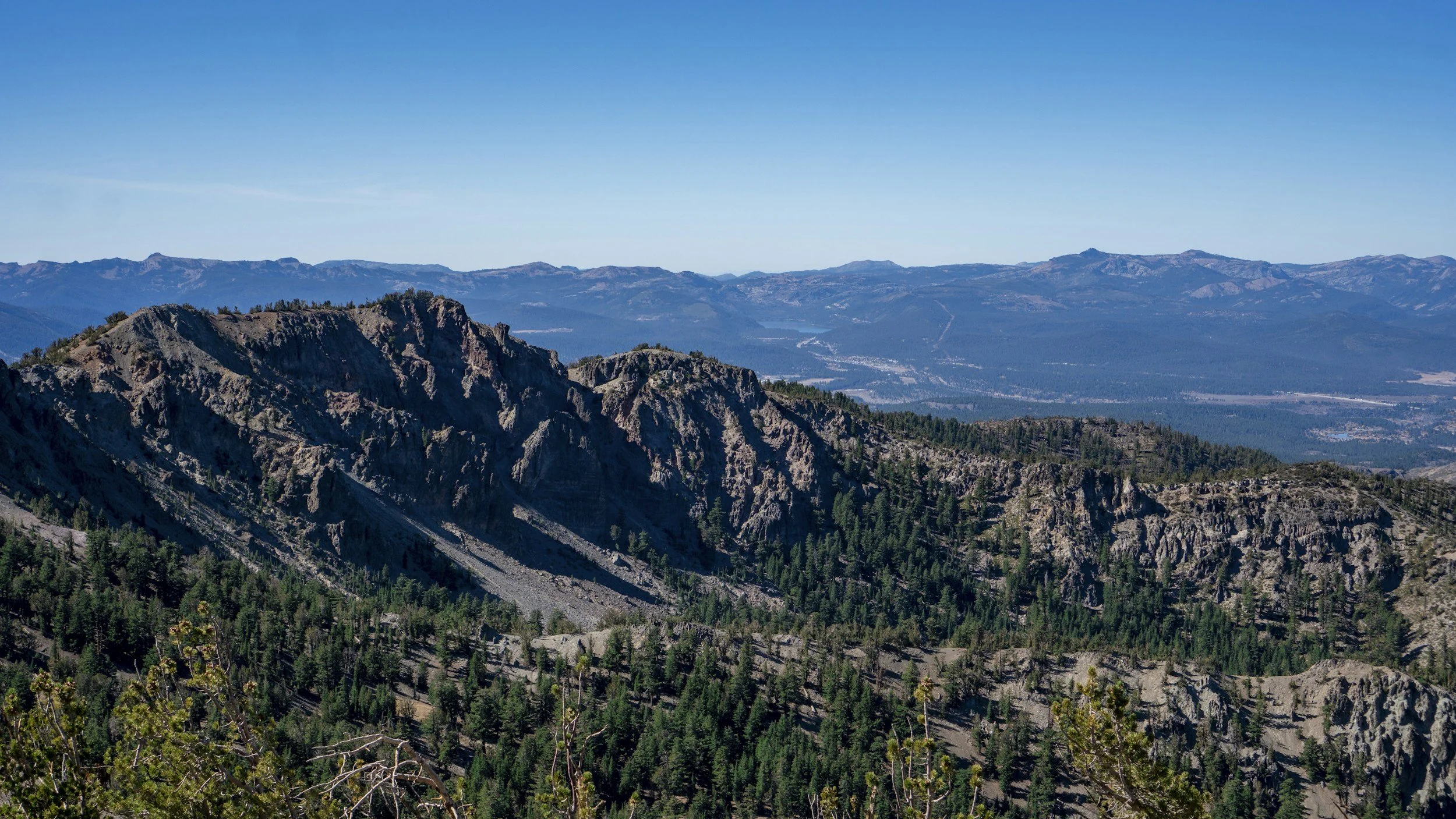 Mountain landscape with rugged peaks, green forested slopes, and a clear blue sky.