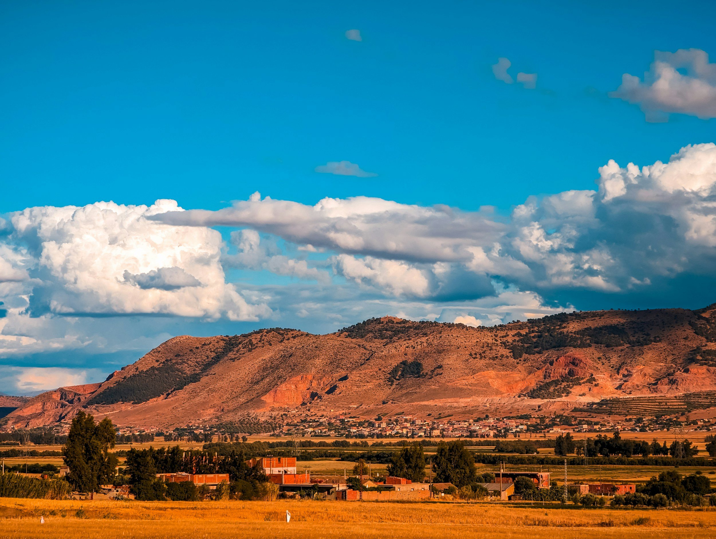A landscape of rolling mountains bathed in warm light, with a blue sky and fluffy white clouds above, and a rural village at the base of the mountains.