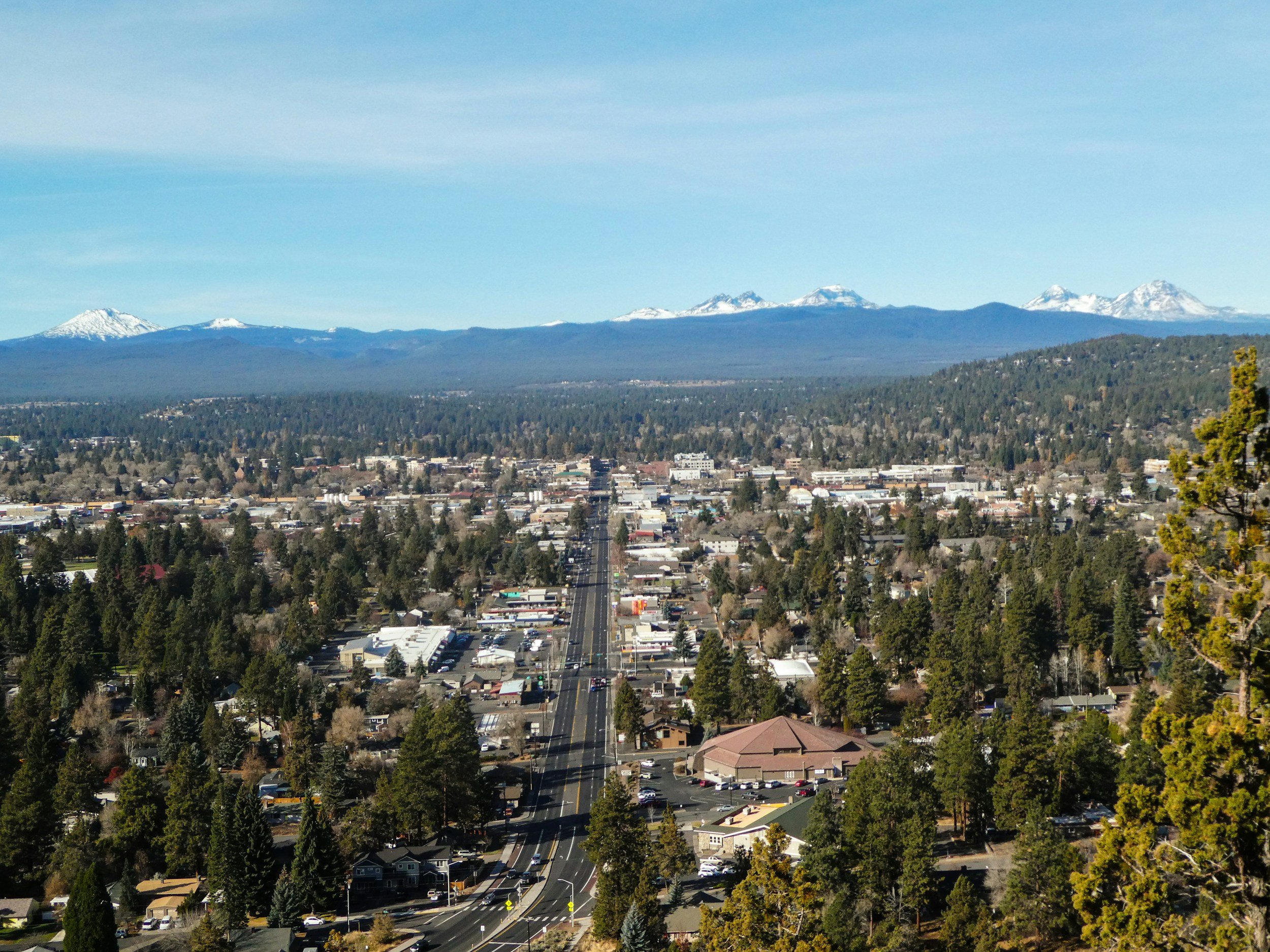 A cityscape with a main road running through it, surrounded by trees, with snowy mountains in the background under a clear blue sky.