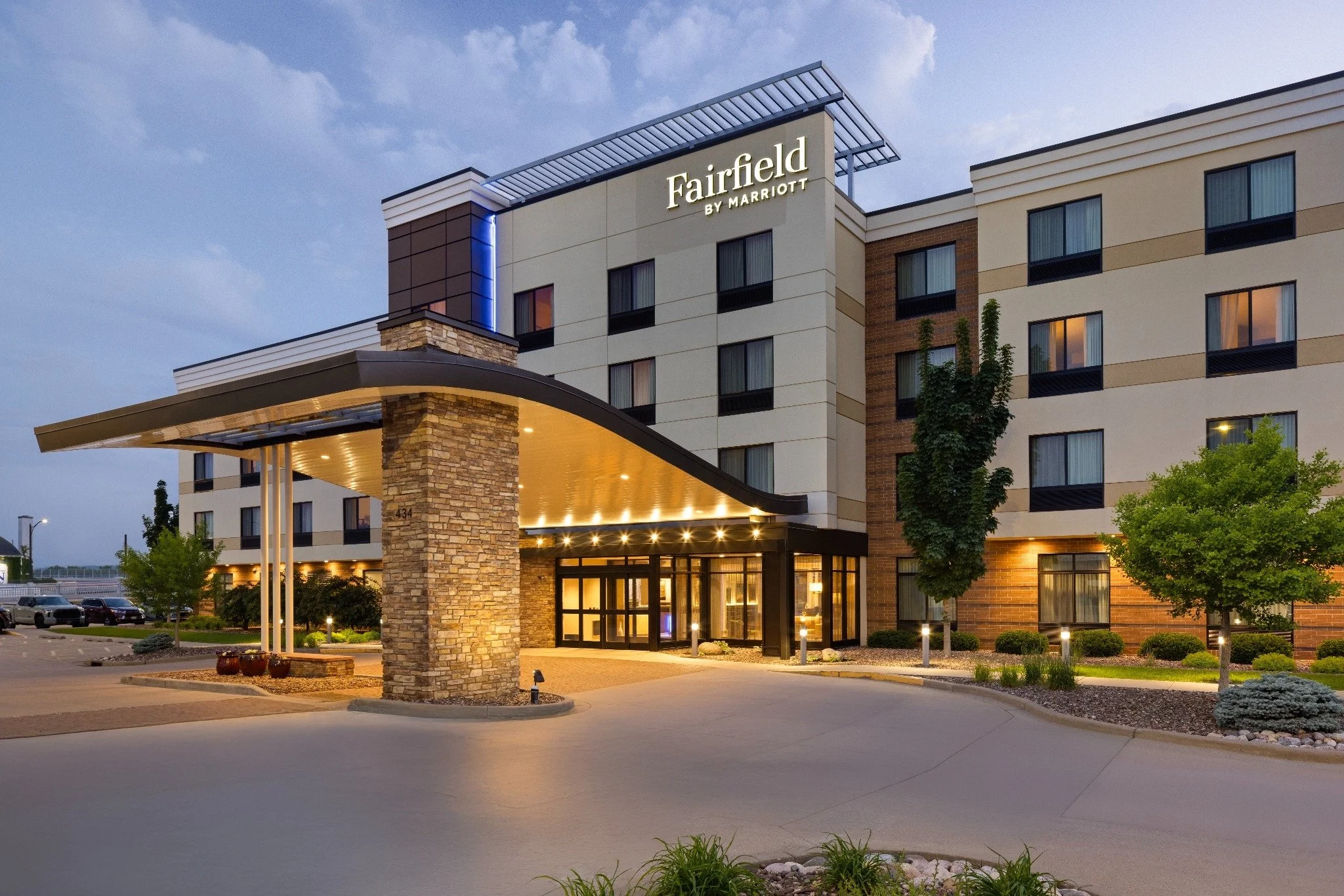 Exterior view of the Fairfield by Marriott hotel at dusk, featuring a modern building with a covered driveway, stone and brick facade, illuminated entrance, and landscaped surroundings with trees and shrubs.
