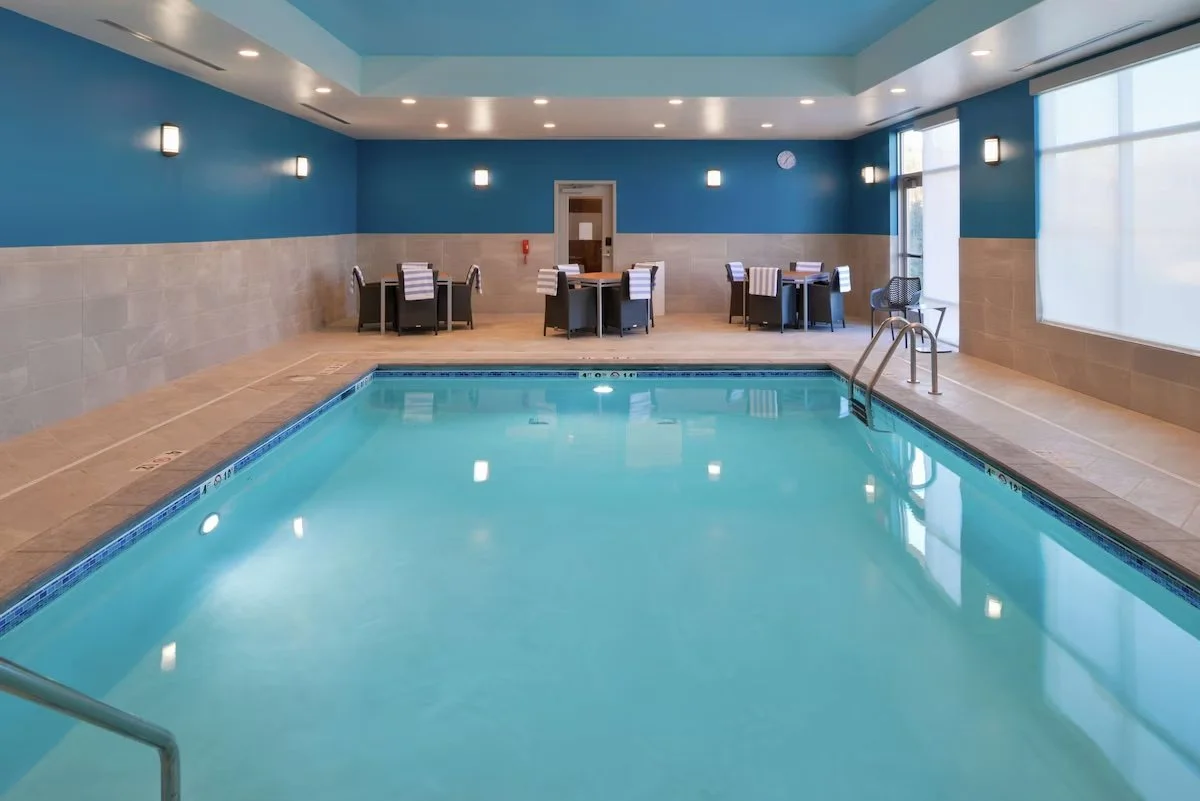 Indoor swimming pool with clear blue water, surrounded by tiled floor and walls, black chairs and tables with striped towels, large windows letting in natural light, and ceiling lights.