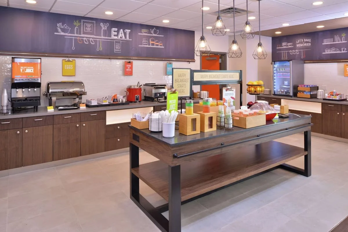 A breakfast buffet area with a table of juice and condiments, and a counter with coffee, tea, and other food items, decorated with chalkboard-style signs and modern hanging lights.