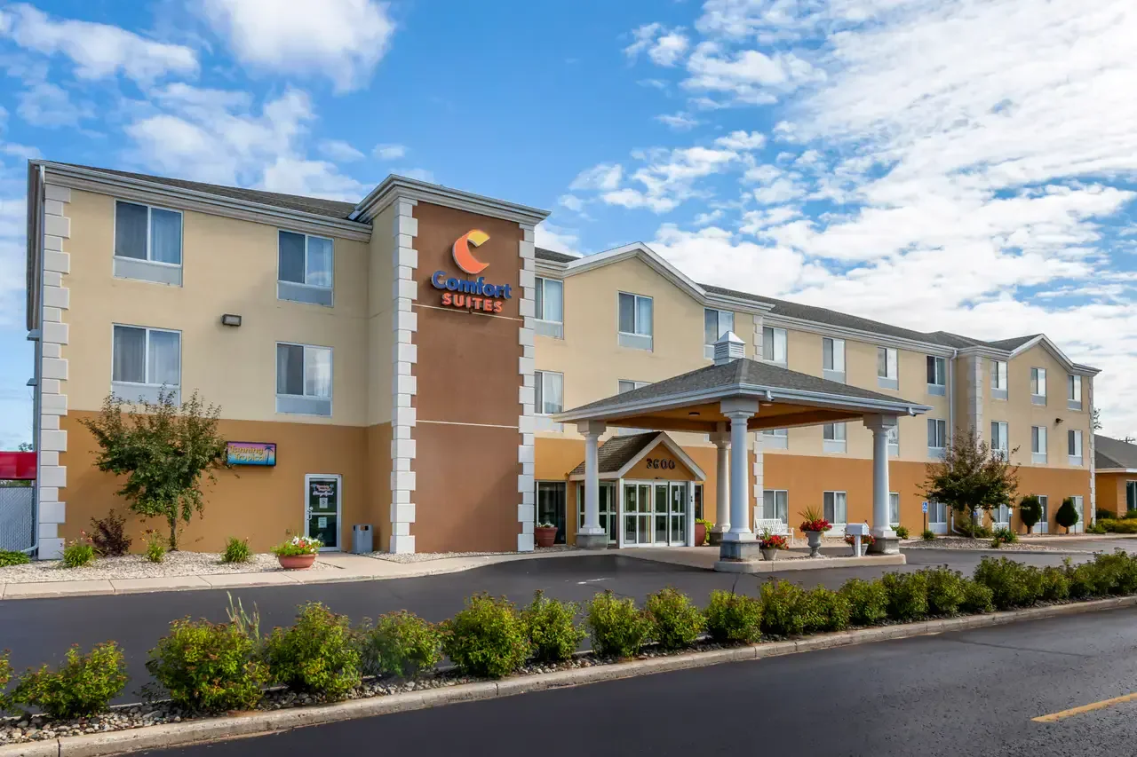 Exterior of a Comfort Suites hotel with a covered entrance, landscaped bushes and trees, under a partly cloudy sky.