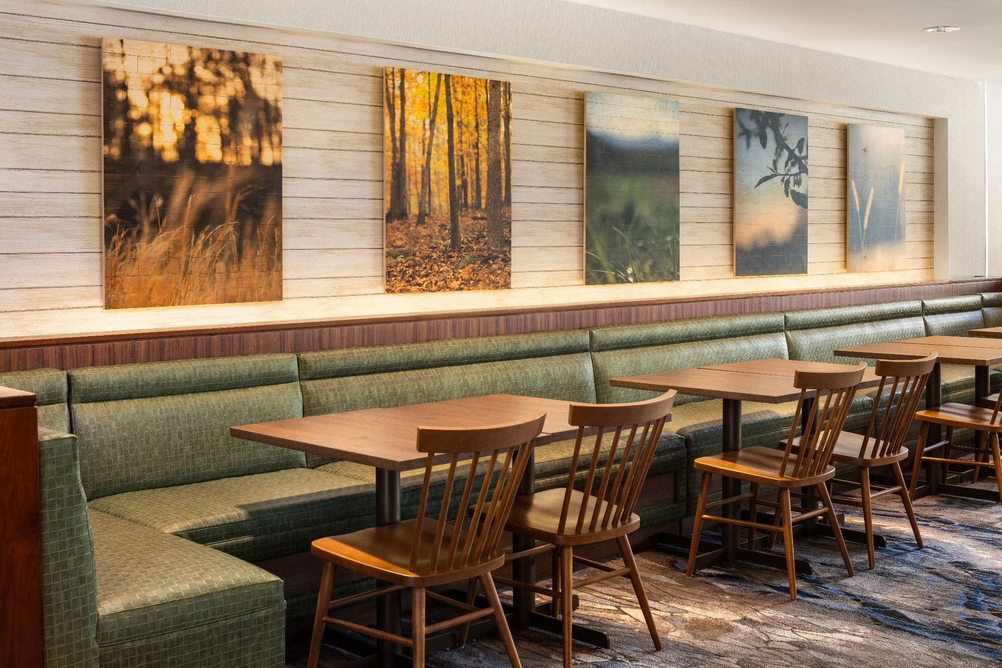 Interior of a restaurant with wooden tables, wooden chairs, and a long green upholstered bench along a wall decorated with five framed landscape photographs of trees and nature scenes.