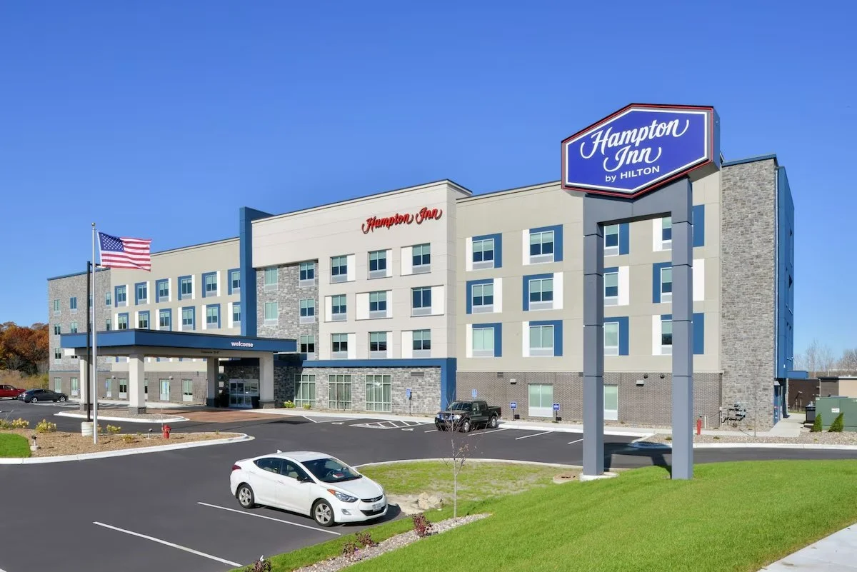 Exterior view of a Hampton Inn hotel with a parking lot, American flags, and a clear blue sky.